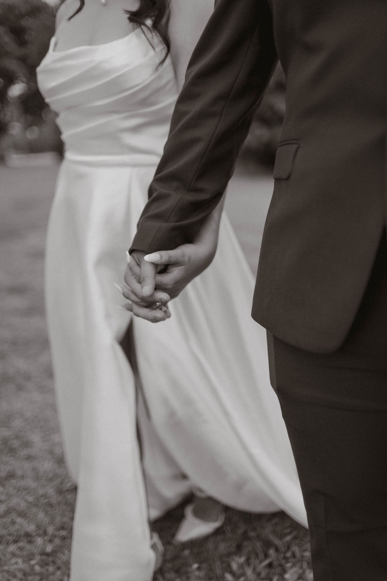 Black and white close-up of bride and groom holding hands, showcasing wedding attire details.