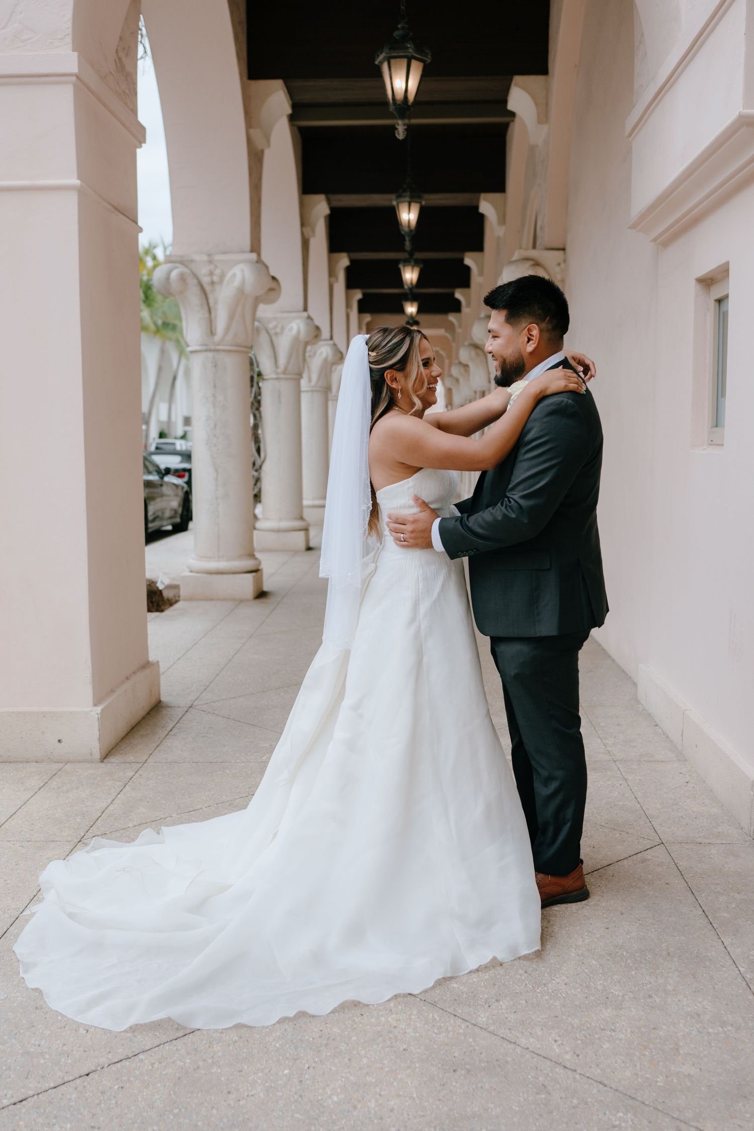 Bride and groom embracing under an elegant archway, bride wearing a long white gown and veil with a dark-suited groom.