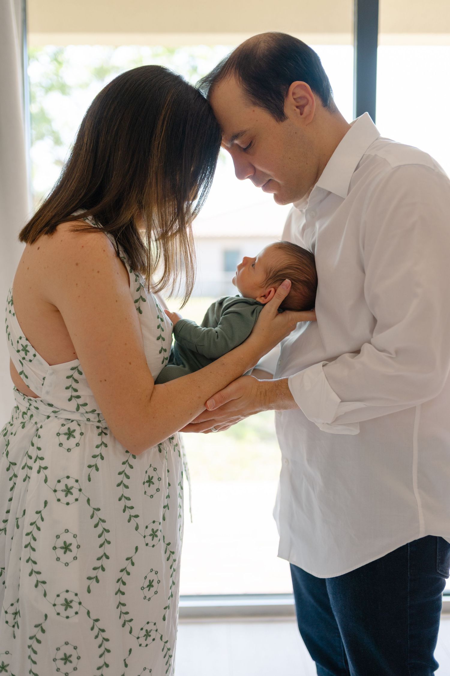 Parents tenderly hold their sleeping newborn baby together near a bright window.