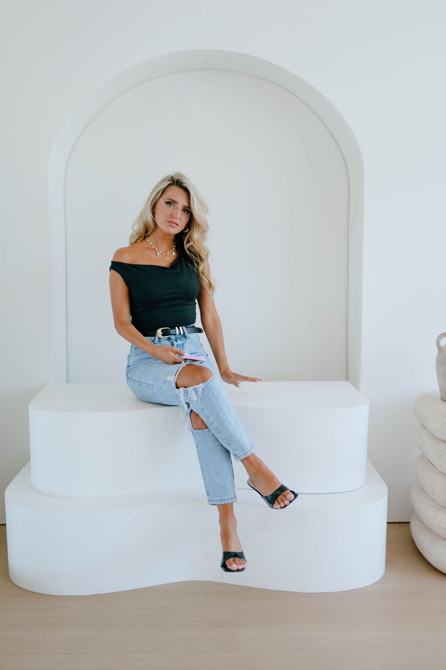 Blonde woman in black off-shoulder top and ripped jeans sitting confidently on a white tiered platform.