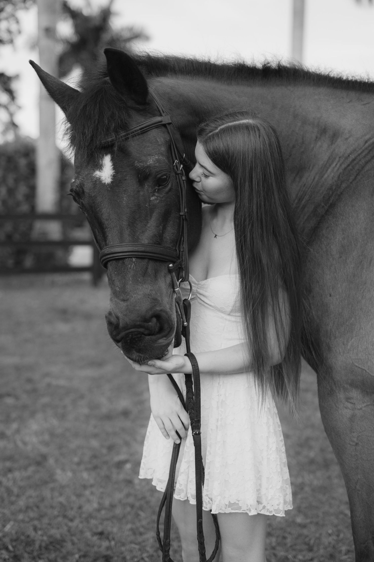 Young woman in white dress tenderly embraces a bay horse in a black and white equestrian portrait.