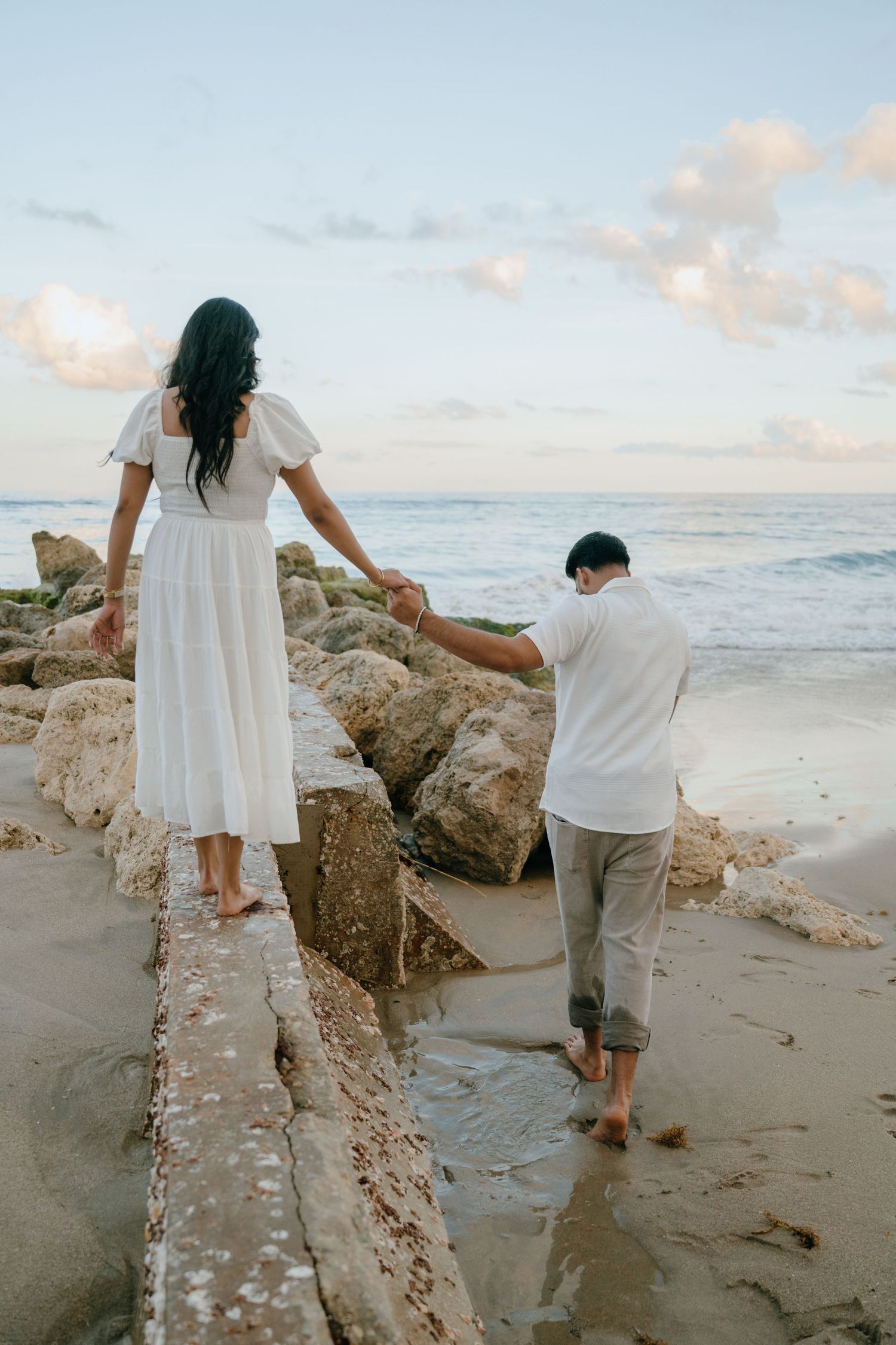 Couple holding hands while walking on rocky beach shoreline at sunset, woman in white dress.
