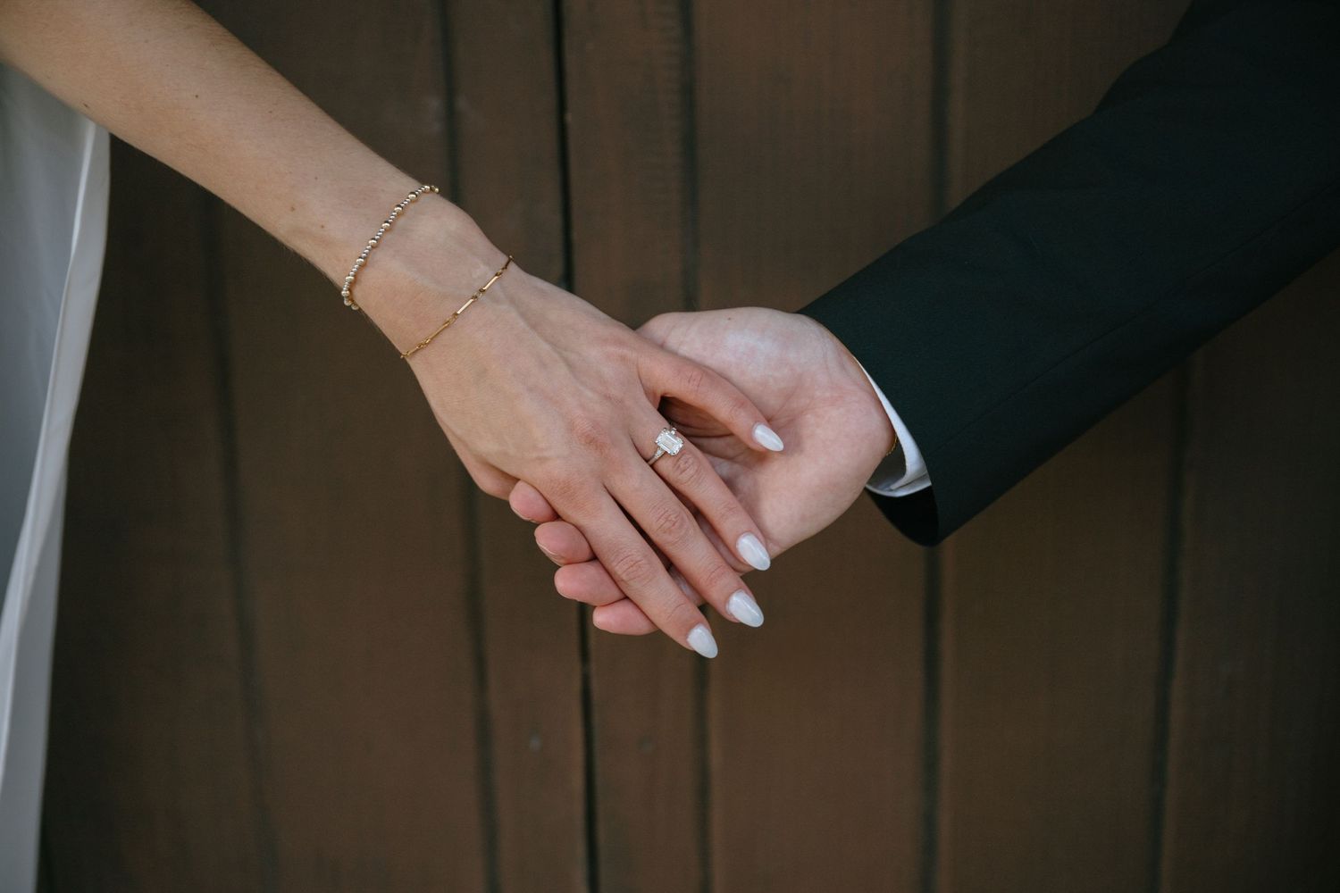 Close-up of bride and groom holding hands, showcasing diamond engagement ring and gold bracelet.