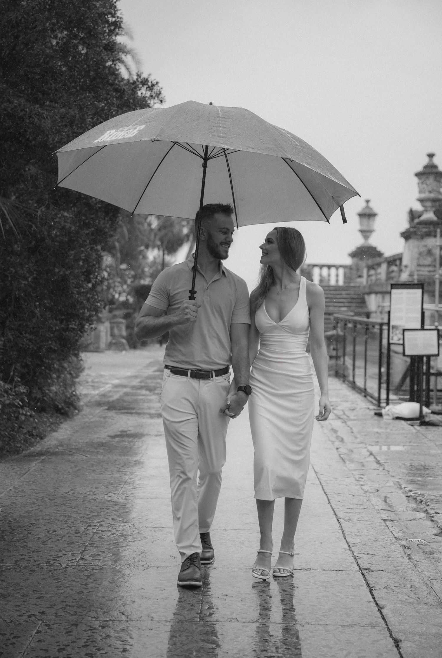Black and white photo of a couple walking hand in hand in the rain under a large umbrella on a wet cobblestone path.