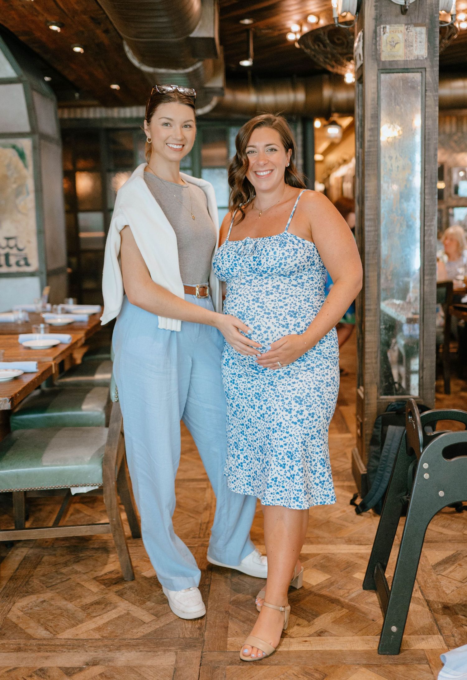 Two women pose together at a baby shower, one in a blue floral dress cradling her pregnant belly.