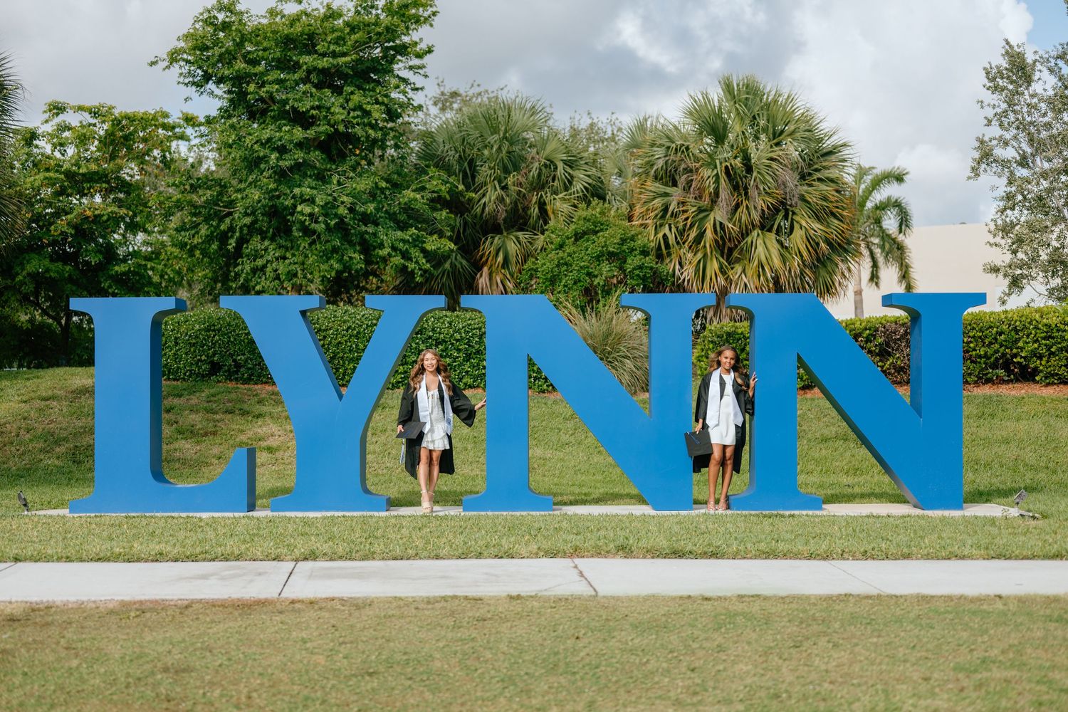 Two graduates in white dresses posing in front of large blue LYNN University letters sign.