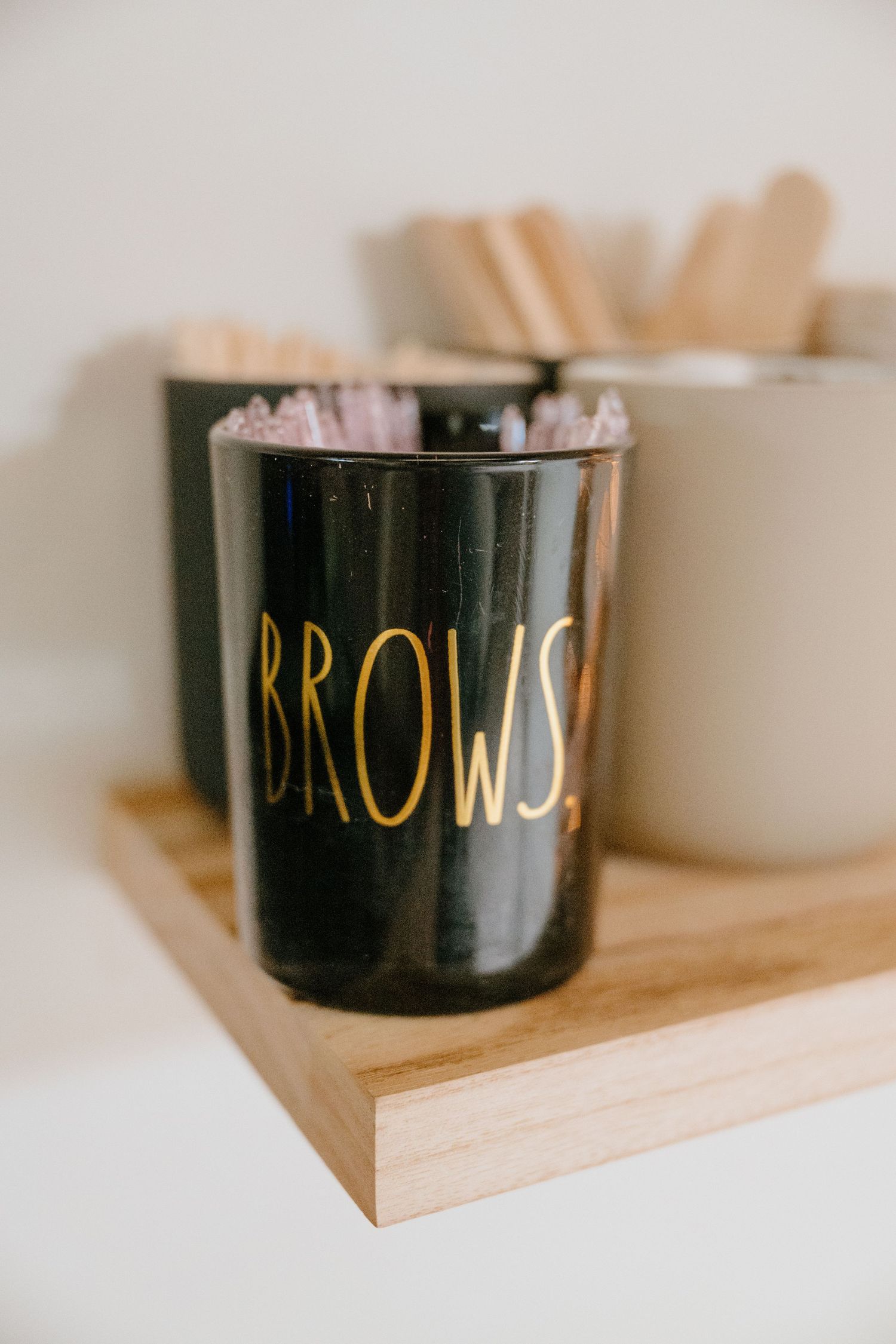 Black Rae Dunn BROWS canister holding waxing sticks on a wooden tray in a beauty salon.