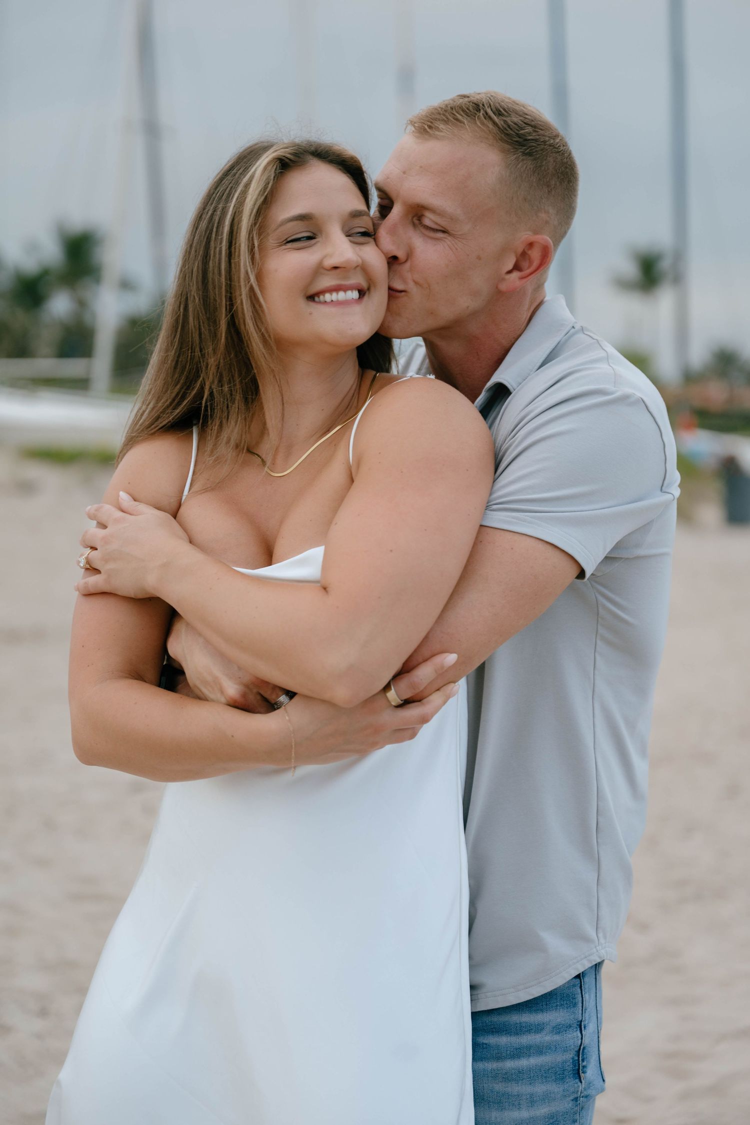 Happy couple embracing on a sandy beach, man kissing woman's cheek as she smiles in a white dress.