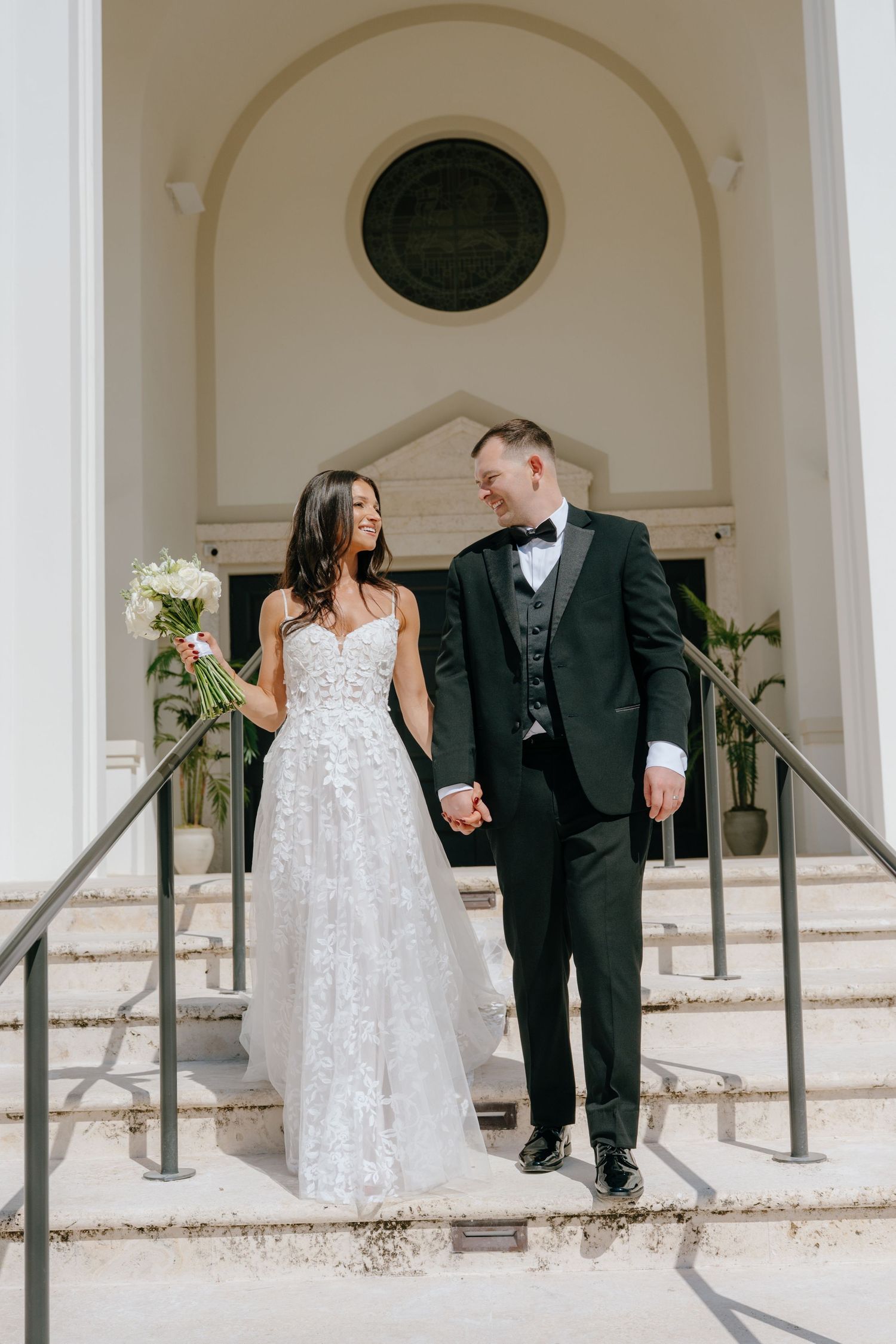 Bride and groom walking down church steps, she holds white bouquet, he wears a black tuxedo.
