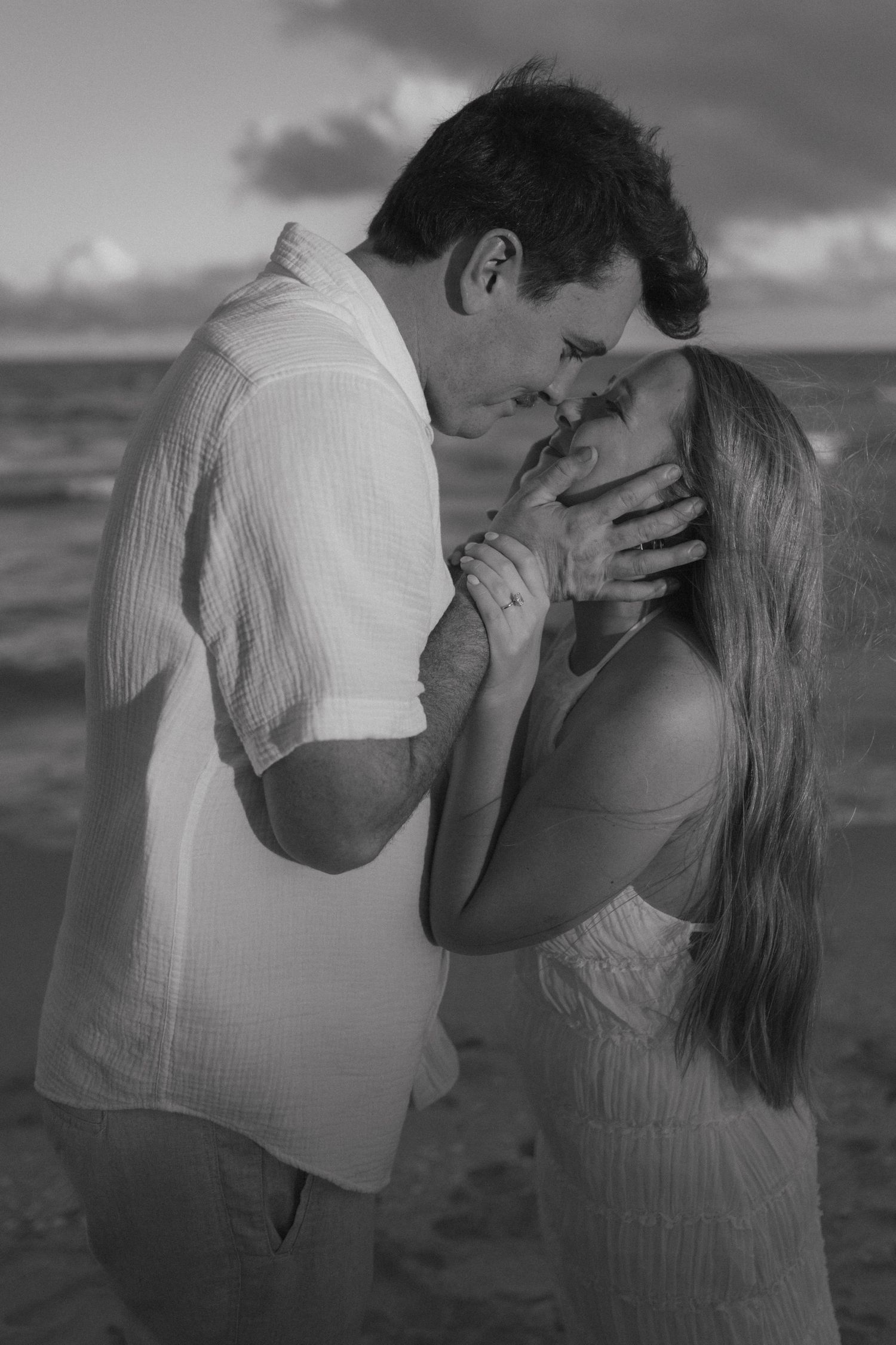 Black and white romantic beach photo of a couple nearly kissing at sunset with ocean waves behind them.