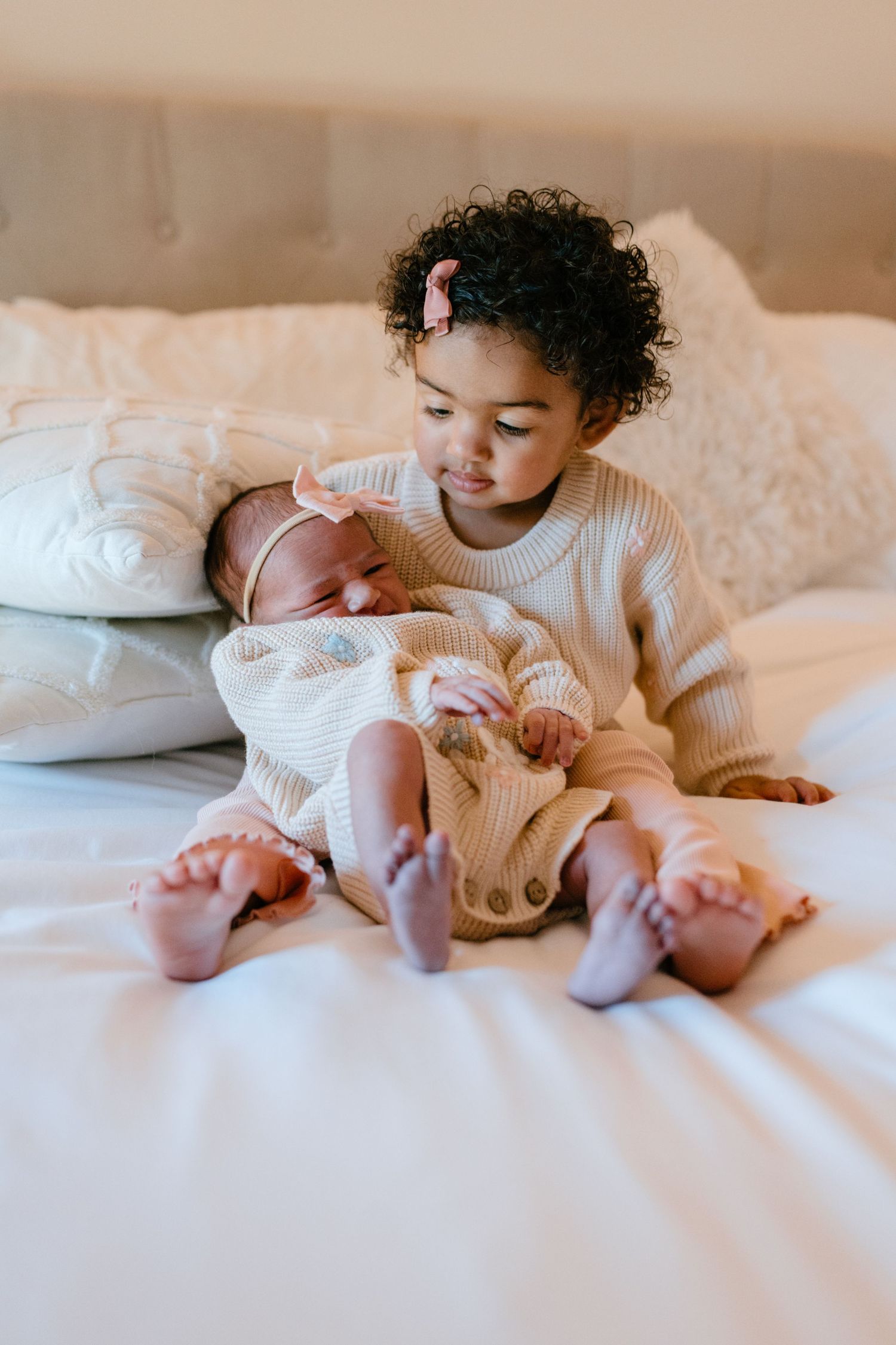 Young toddler girl in cream sweater gently holding newborn sibling on white bed.