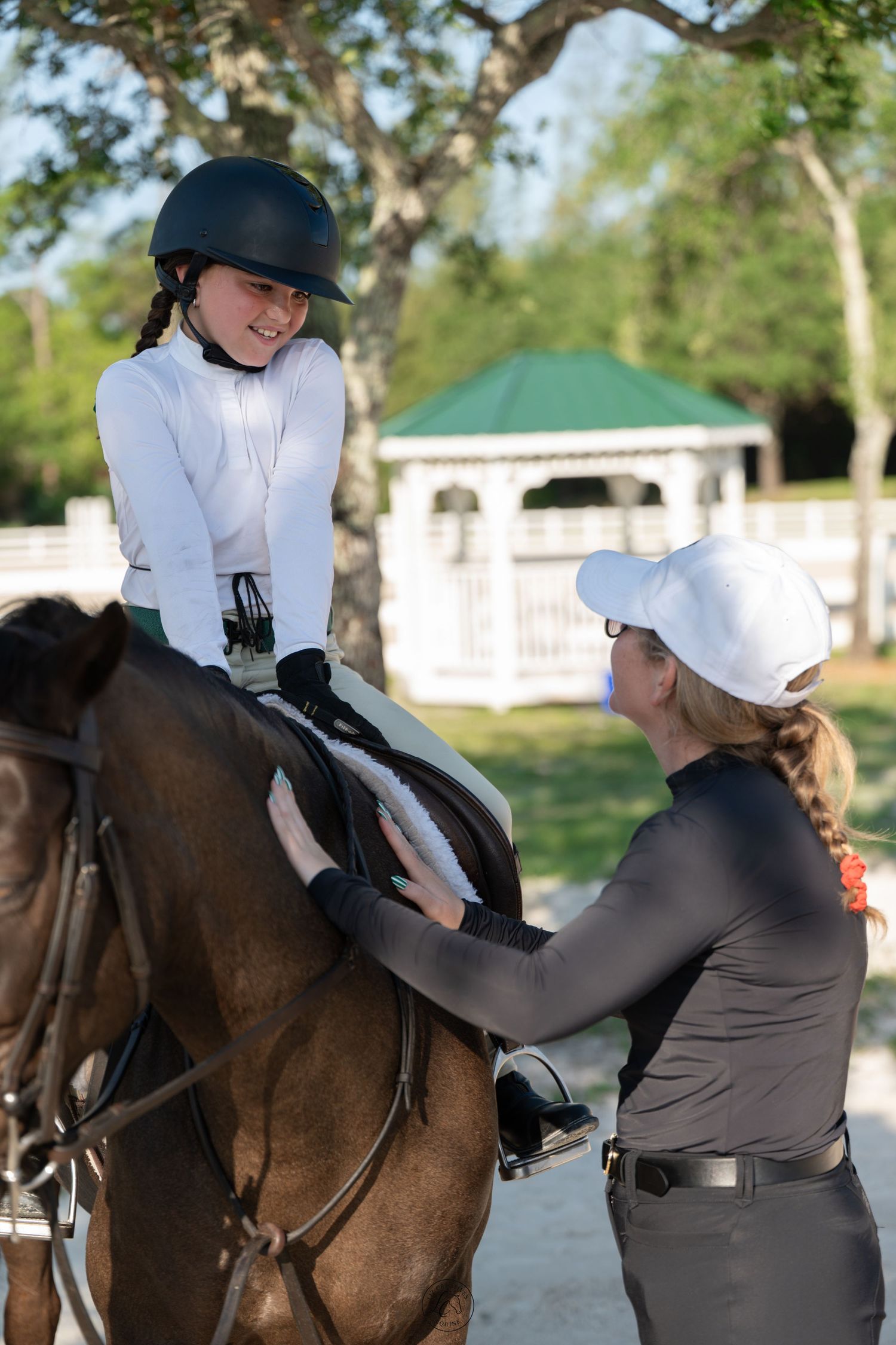 Smiling young equestrian on horseback receiving saddle adjustment from instructor outdoors.