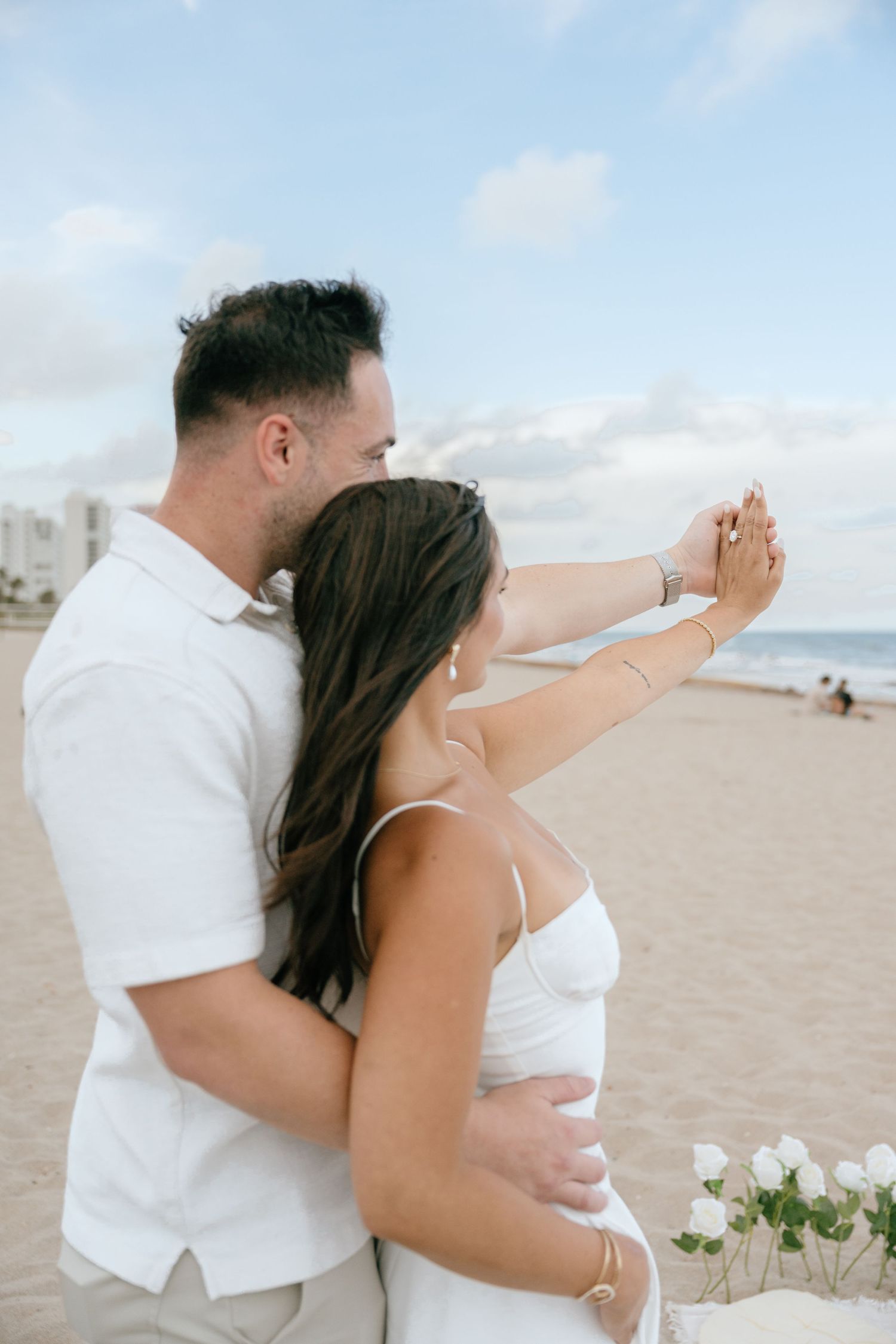 Newly engaged couple holding hands and admiring ring on sunny beach with ocean in background.