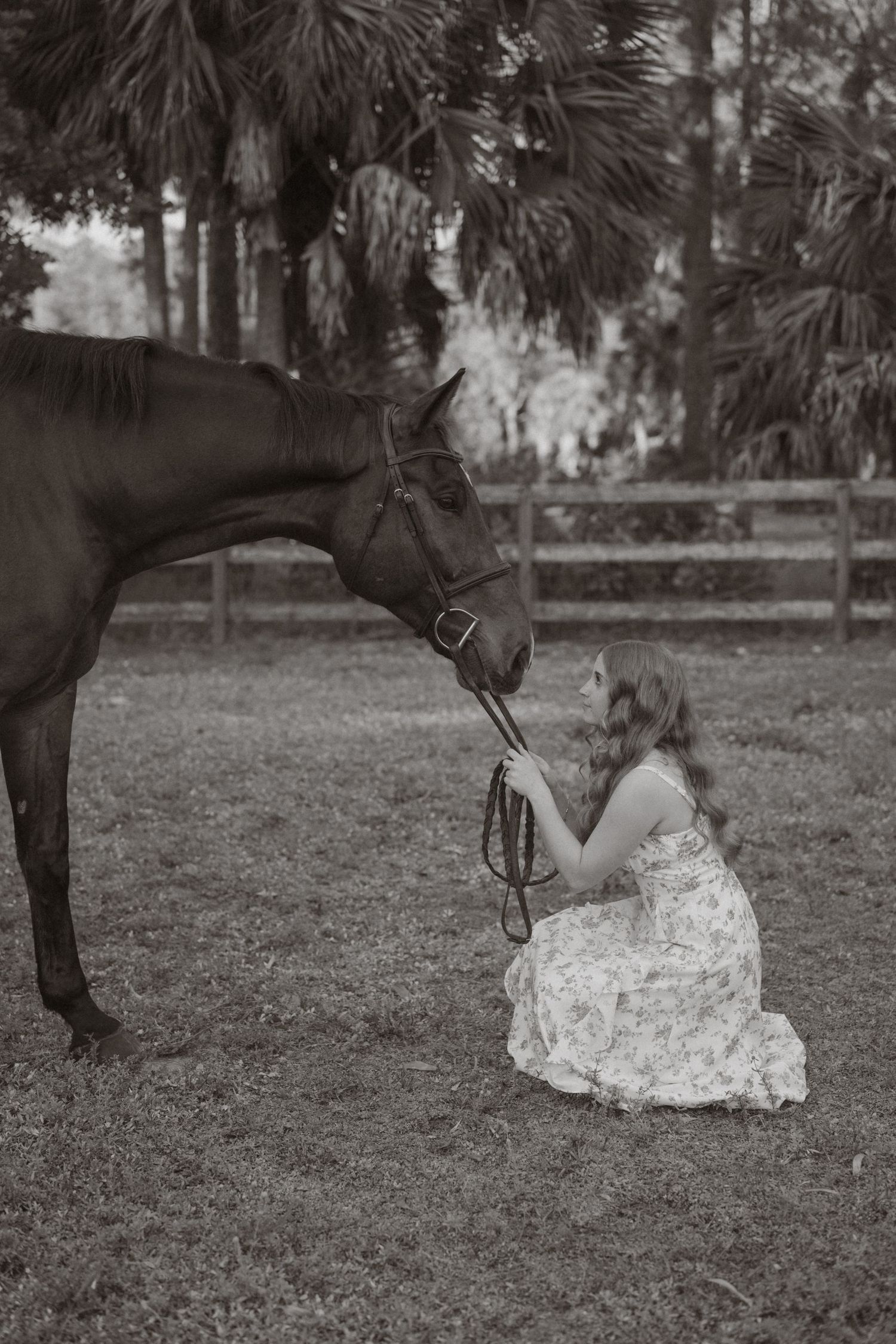 Sepia toned photo of girl in floral dress kneeling before dark bay horse on grass.