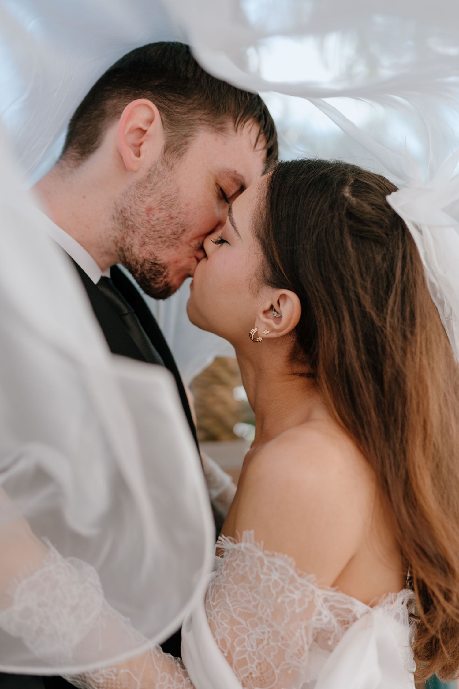 A bride and groom share a romantic kiss under a flowing white veil on their wedding day.