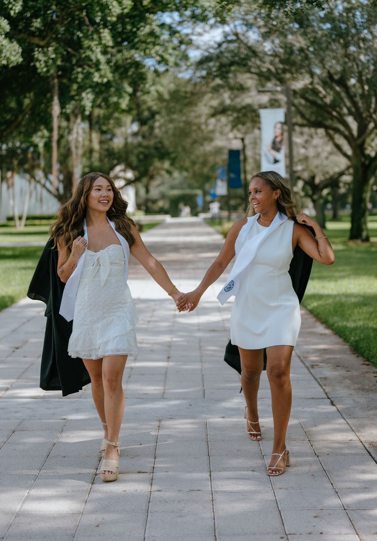 Two female graduates in white dresses holding hands and walking together on a tree-lined campus path.