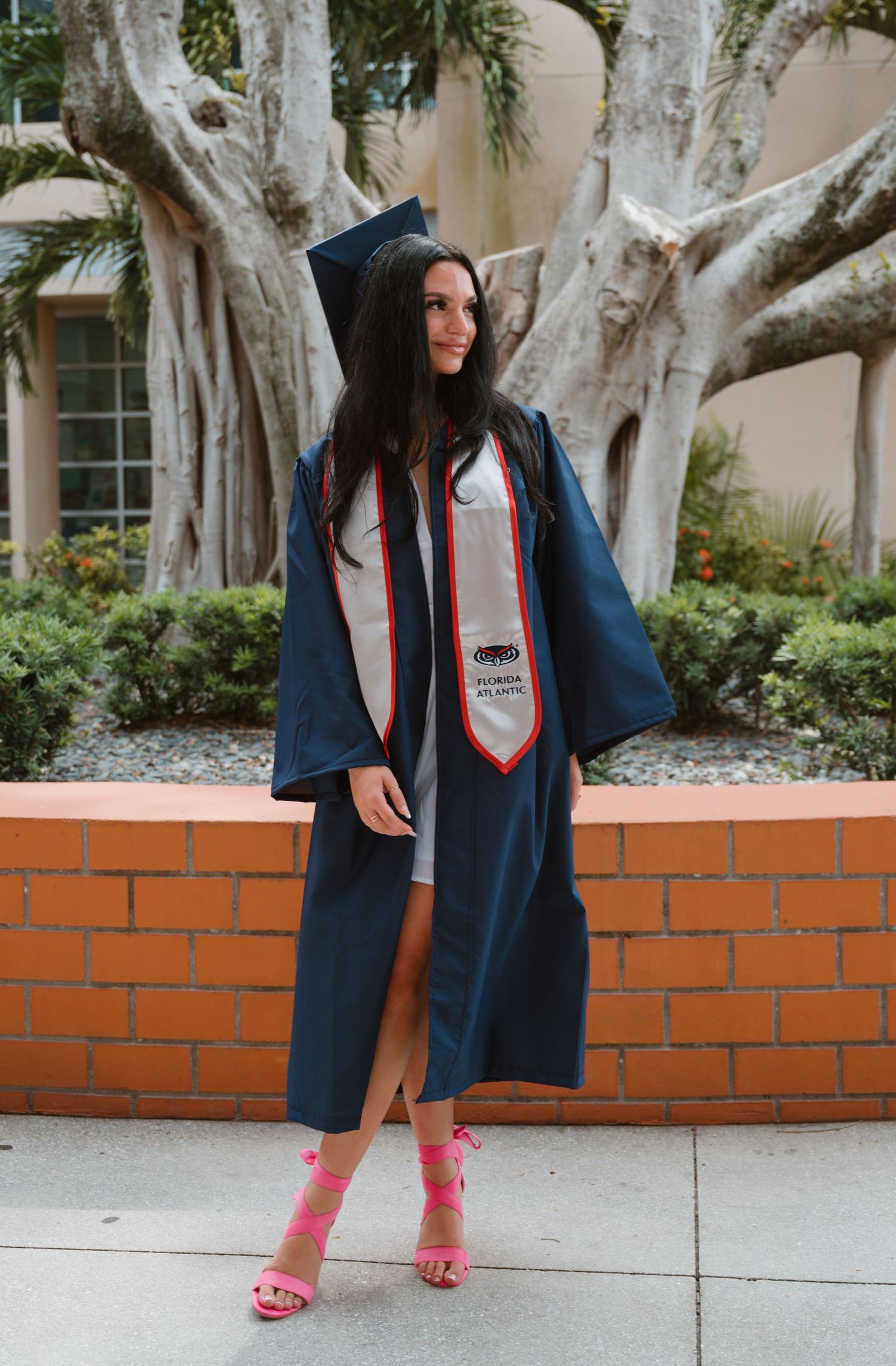 Female graduate in navy FAU cap and gown poses before lush tropical trees in pink heels.