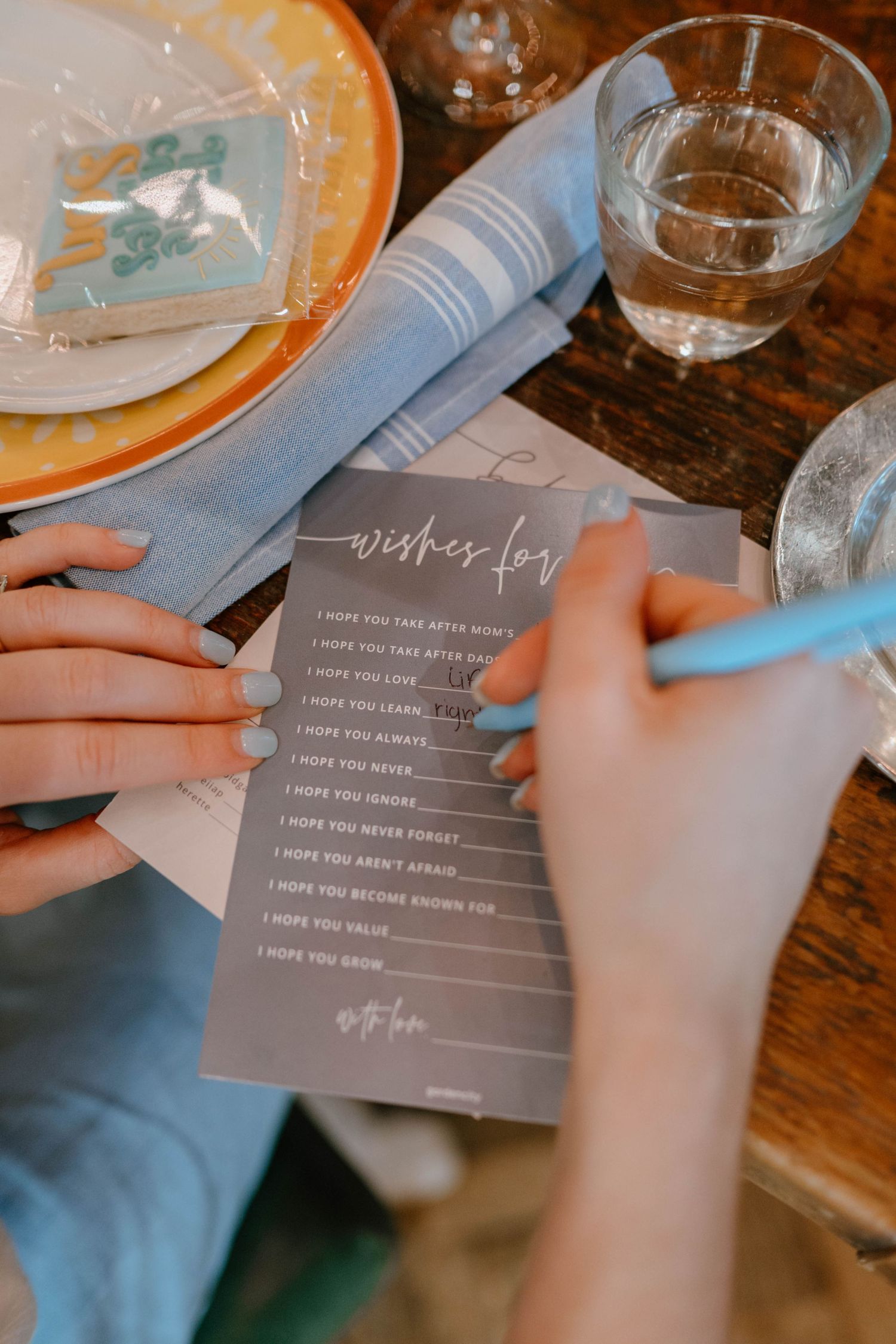 Hands writing wishes on a baby shower card at a decorated table with blue napkins and glassware.