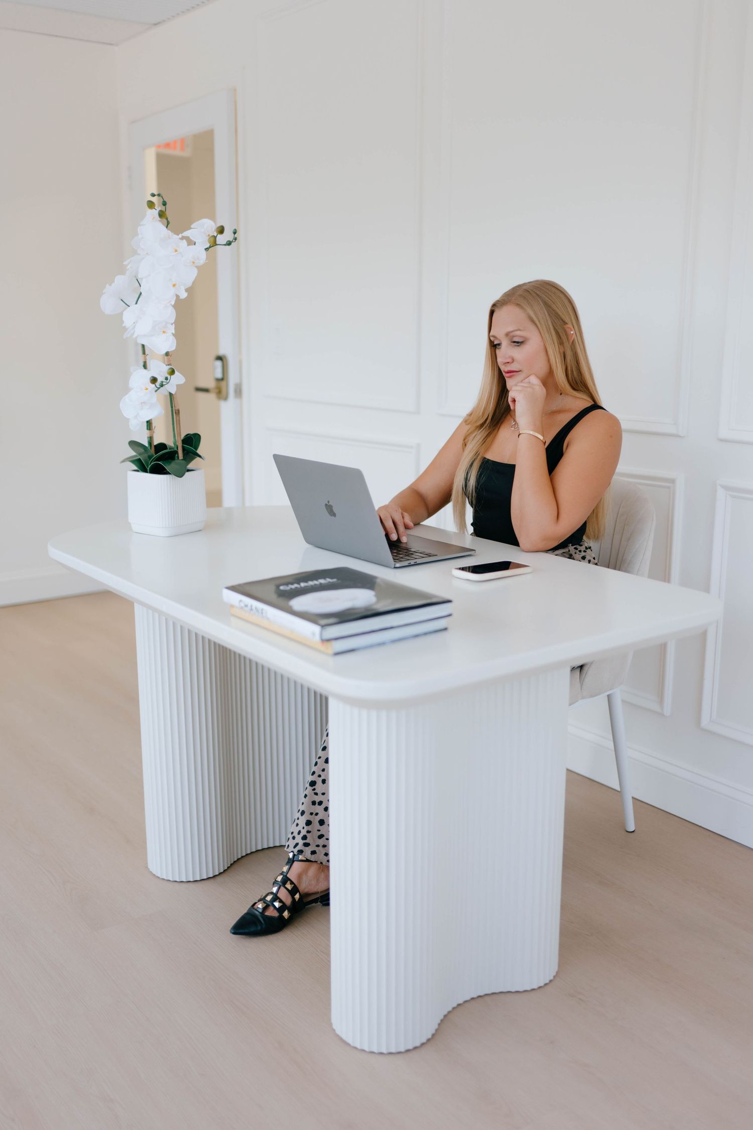 Blonde woman working on a MacBook laptop at a modern white fluted desk in a bright, elegant office space.