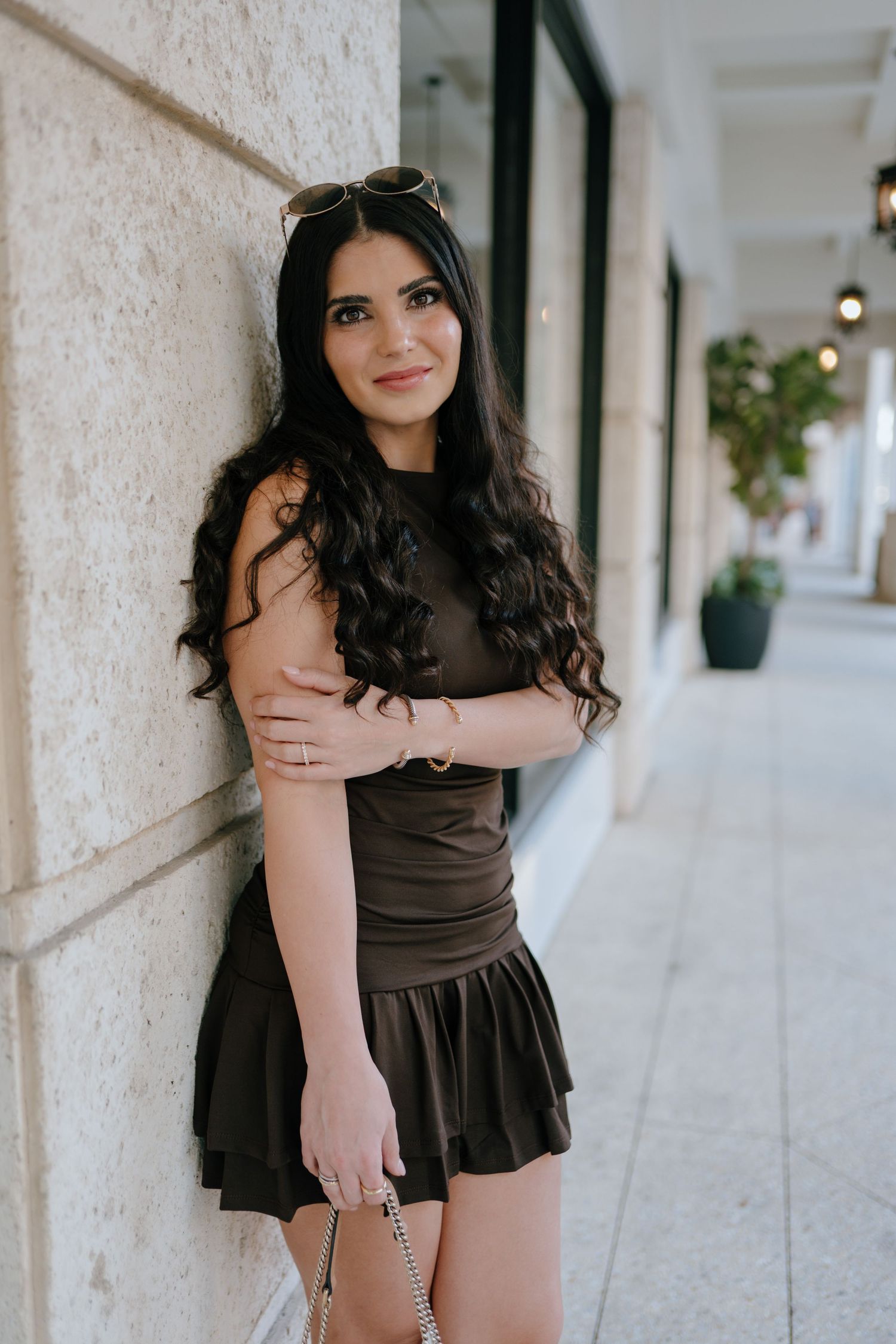 Young woman in dark olive mini dress leaning against stone wall in outdoor corridor.