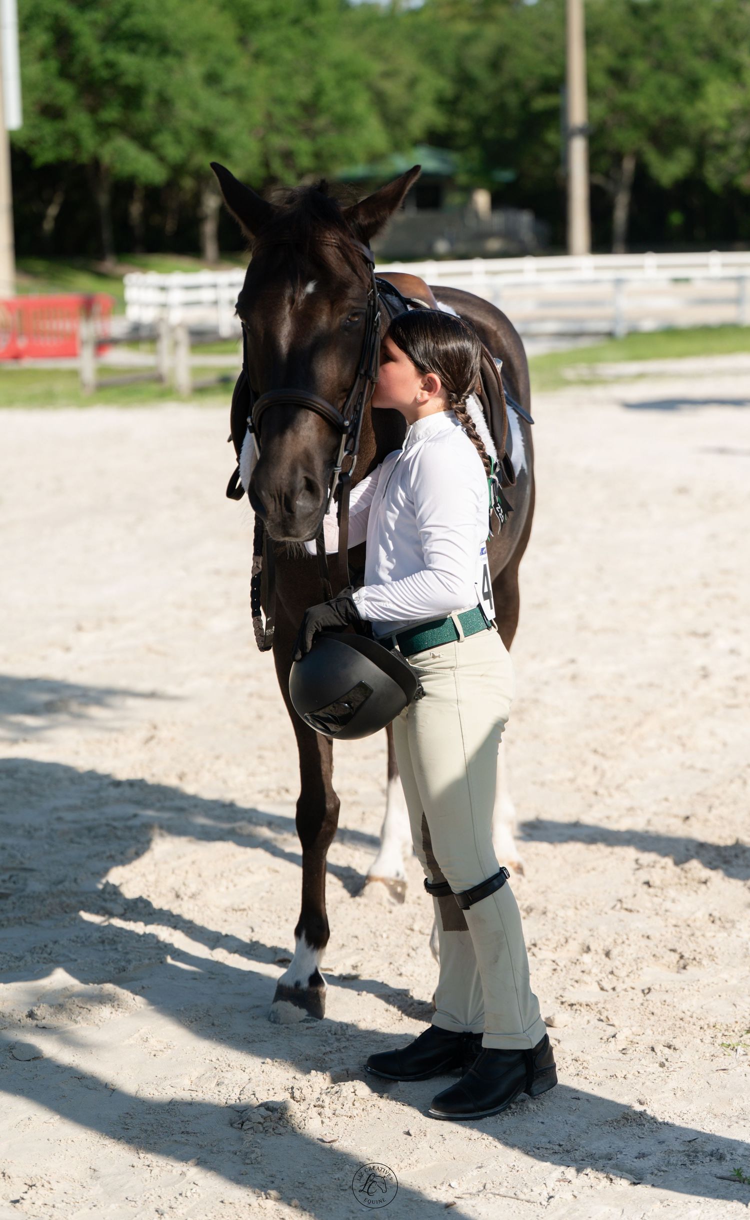 Young equestrian in riding gear sharing tender moment with dark horse in sandy arena.