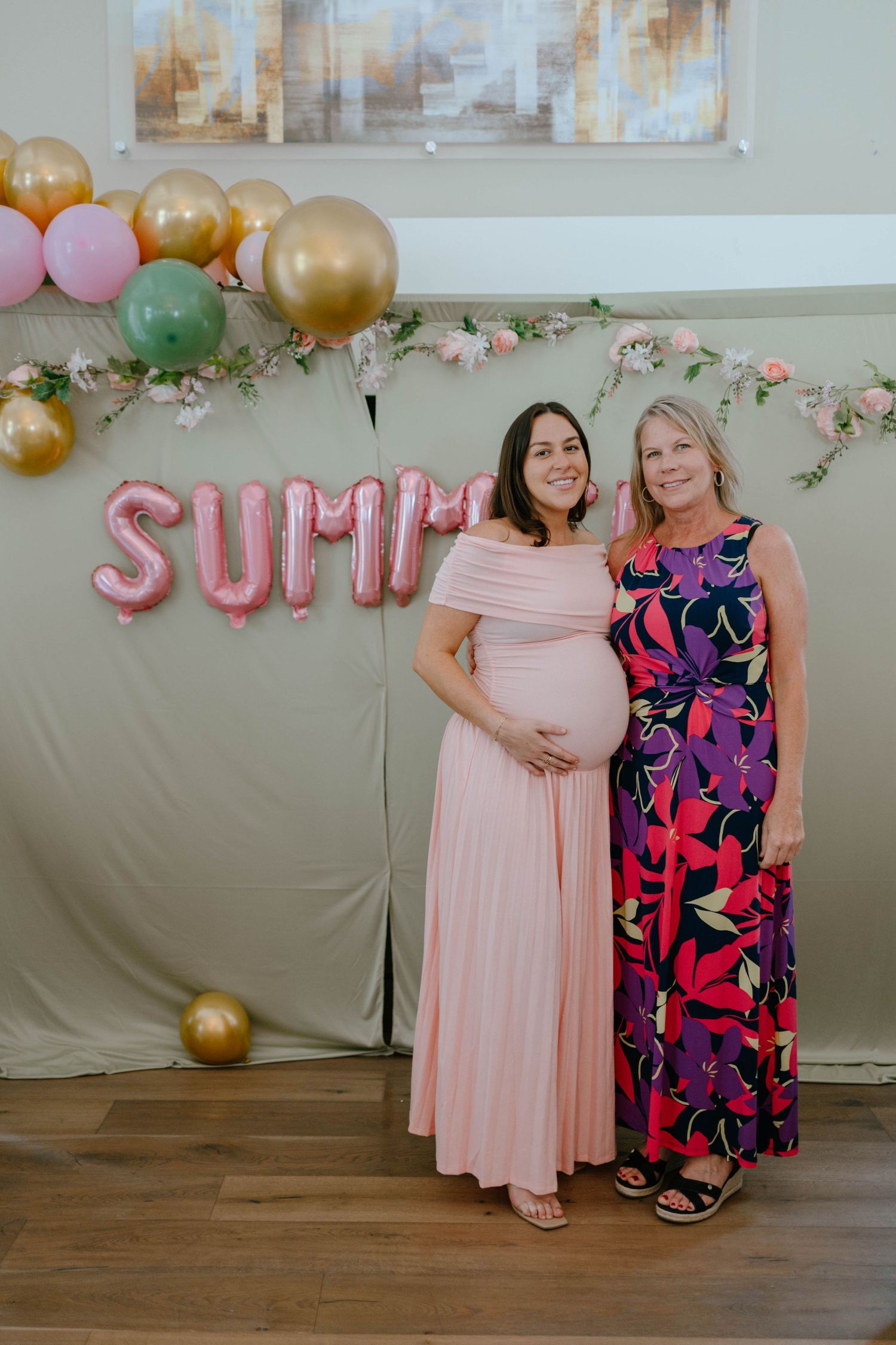 Pregnant woman in pink dress poses with older woman at baby shower with Summer balloon backdrop.