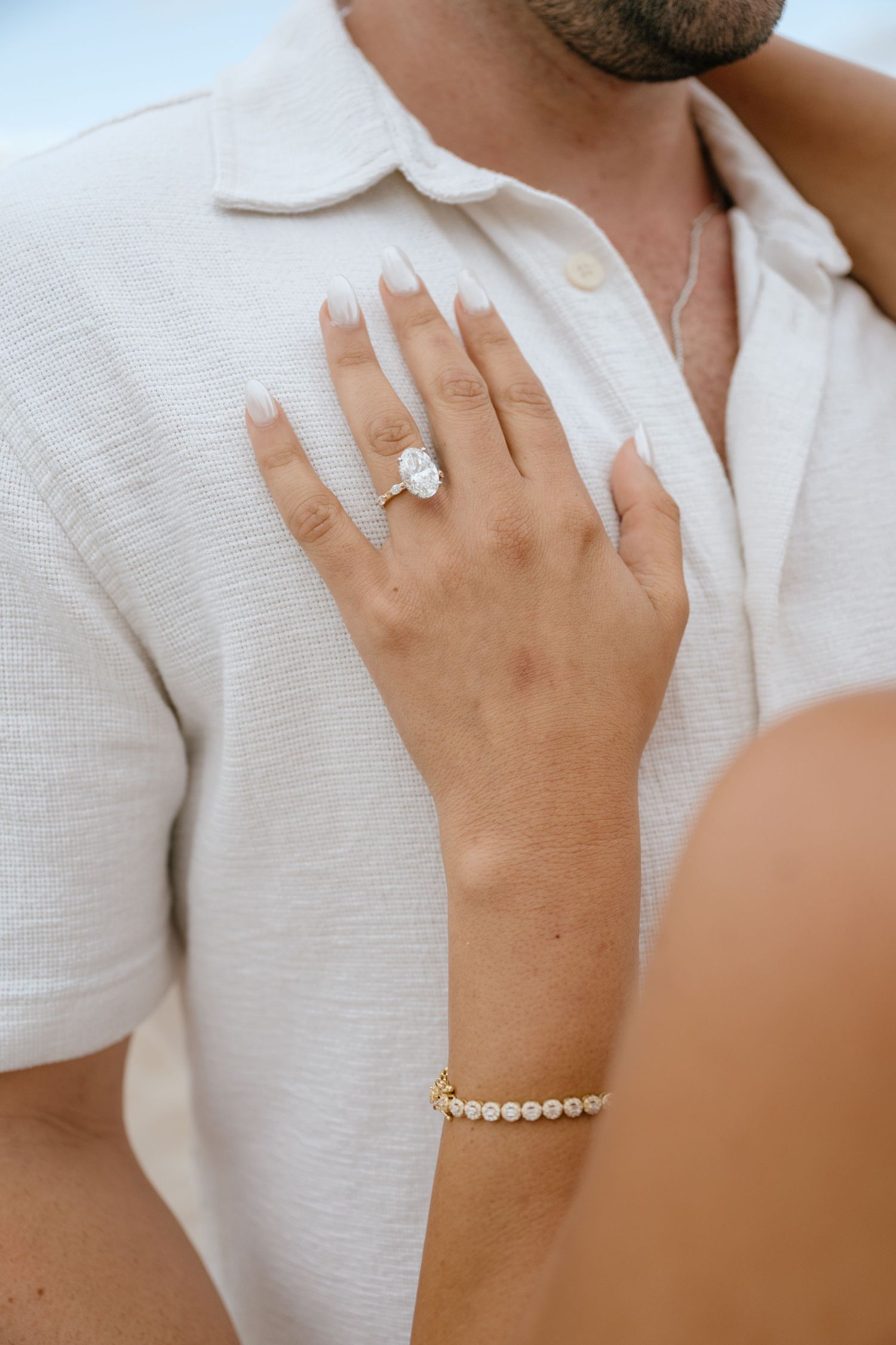 Close-up of oval diamond engagement ring and gold bracelet on womans hand against mans white shirt.