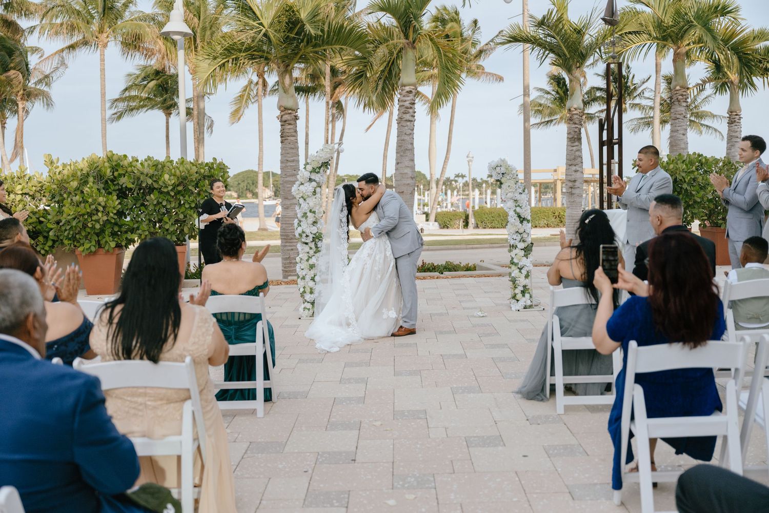 Bride and groom share first kiss as newlyweds at outdoor beach wedding ceremony with palm trees.