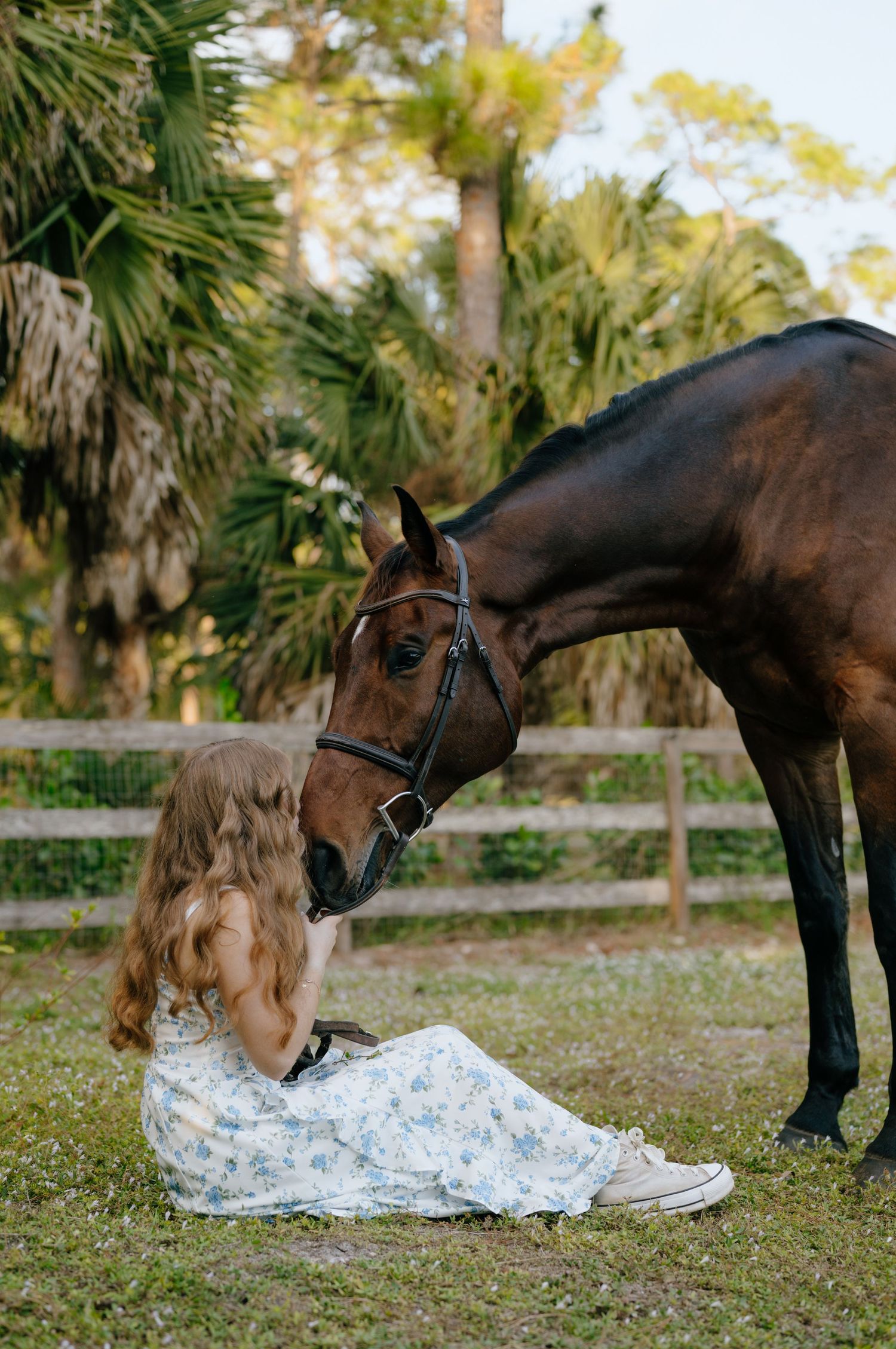 Young woman in floral dress sits on grass sharing tender moment with bay horse.