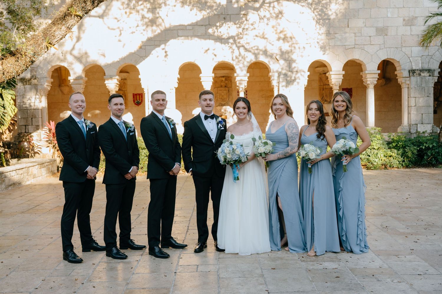 Wedding party of seven posing in courtyard with arched stone colonnade, bridesmaids in dusty blue gowns.