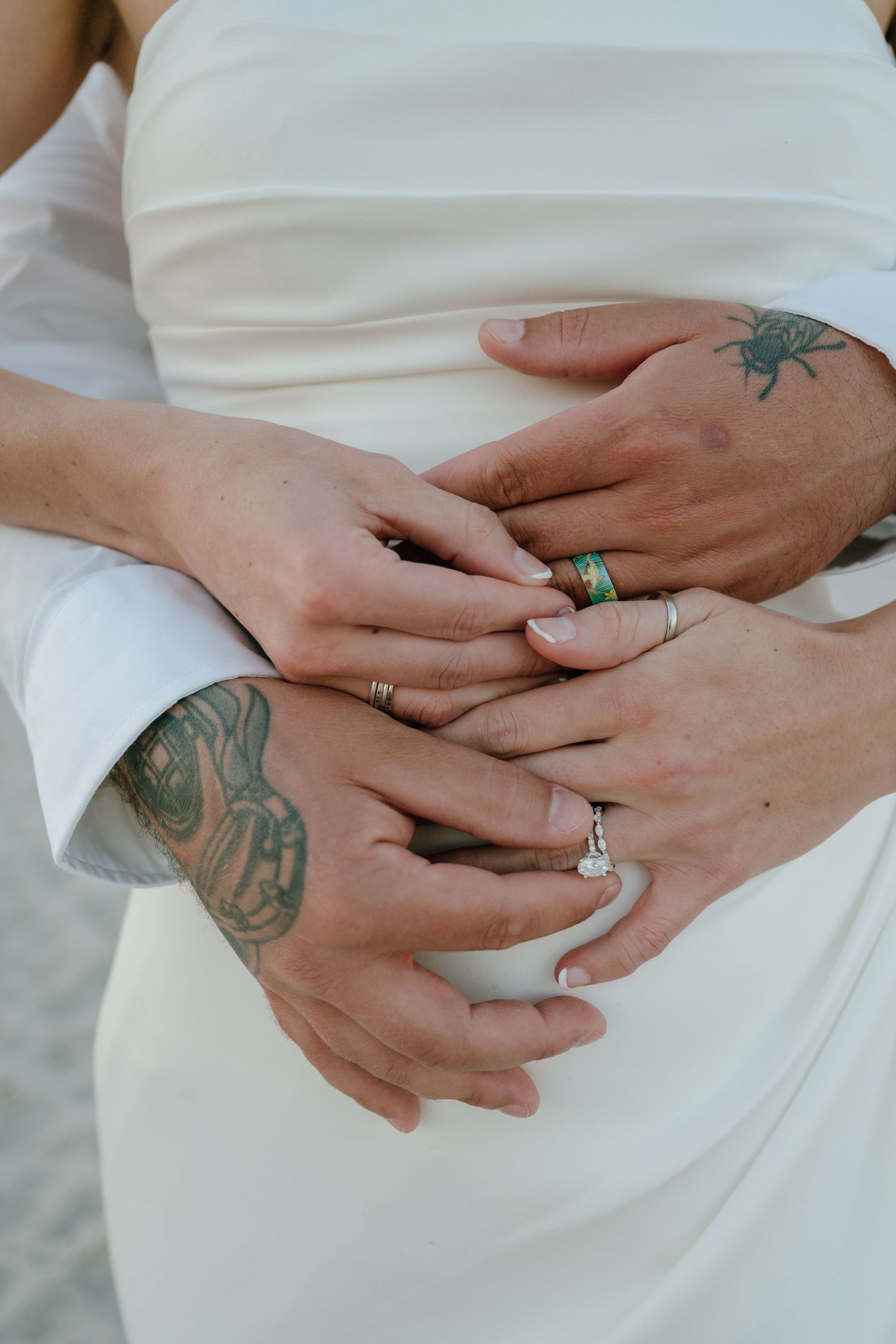 Close-up of bride and groom hands with wedding rings and tattoos at beach wedding.