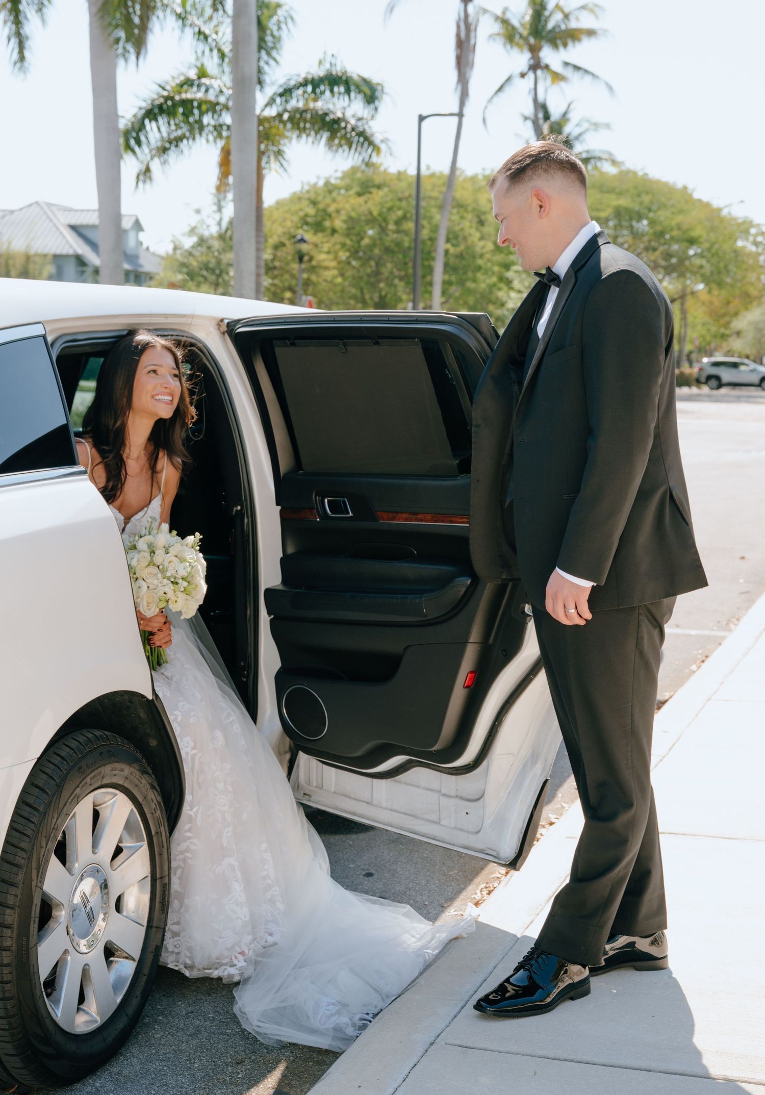 Groom in black tuxedo opening a white SUV door for his smiling bride holding a white bouquet.