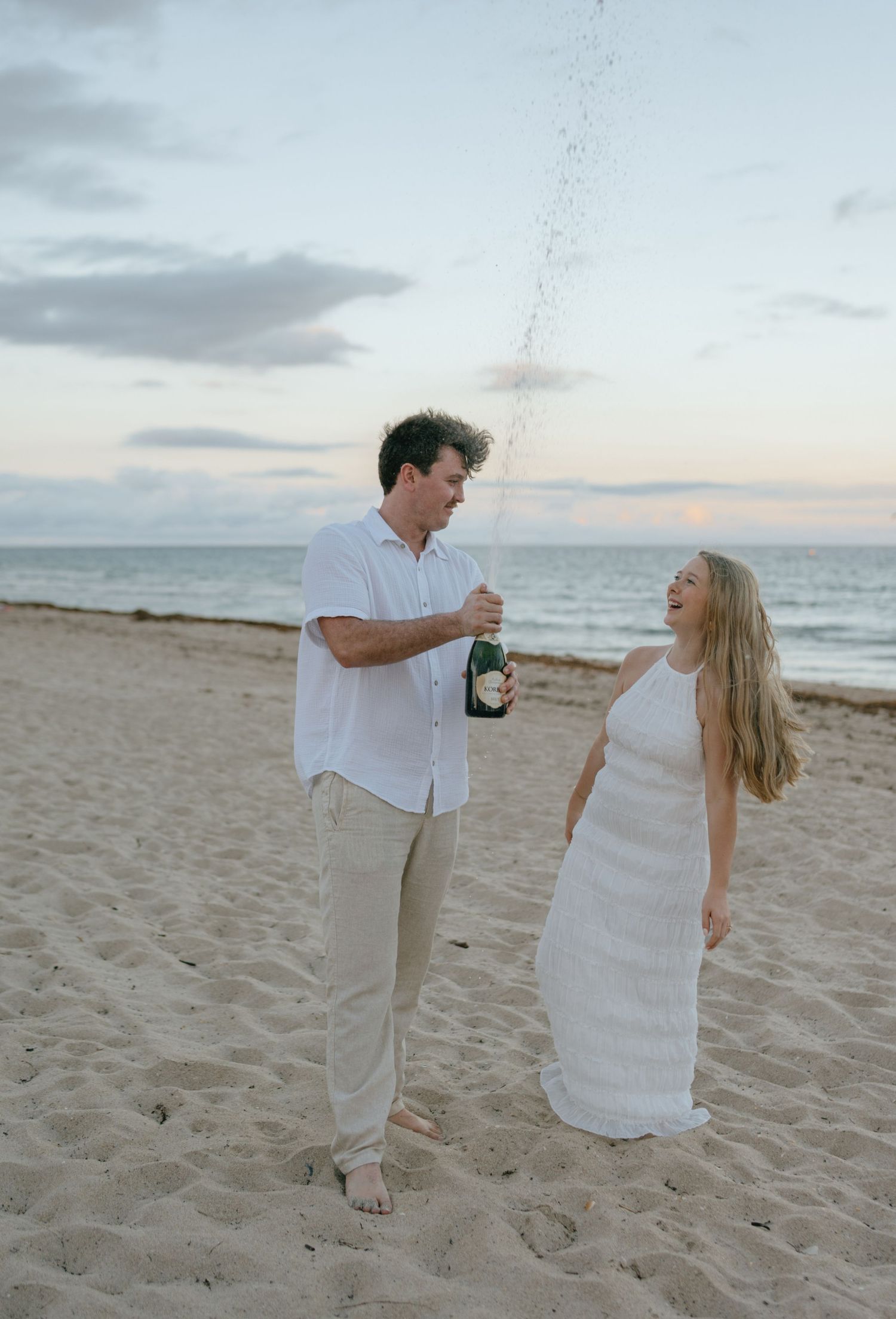Engaged couple celebrating on the beach at sunset as man pops a champagne bottle near the ocean shore.
