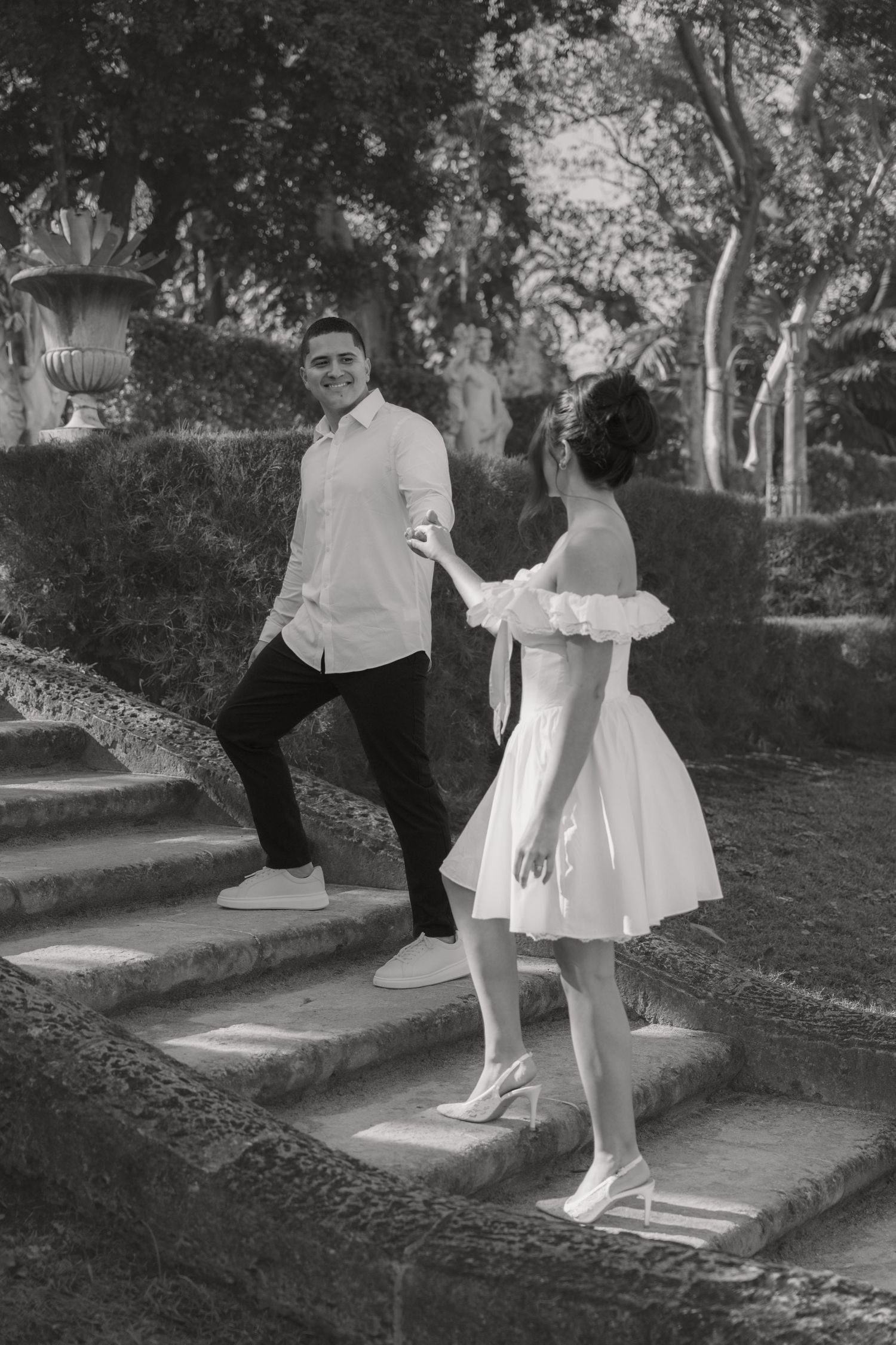 Black and white photo of smiling couple holding hands on stone garden steps, woman in white dress.