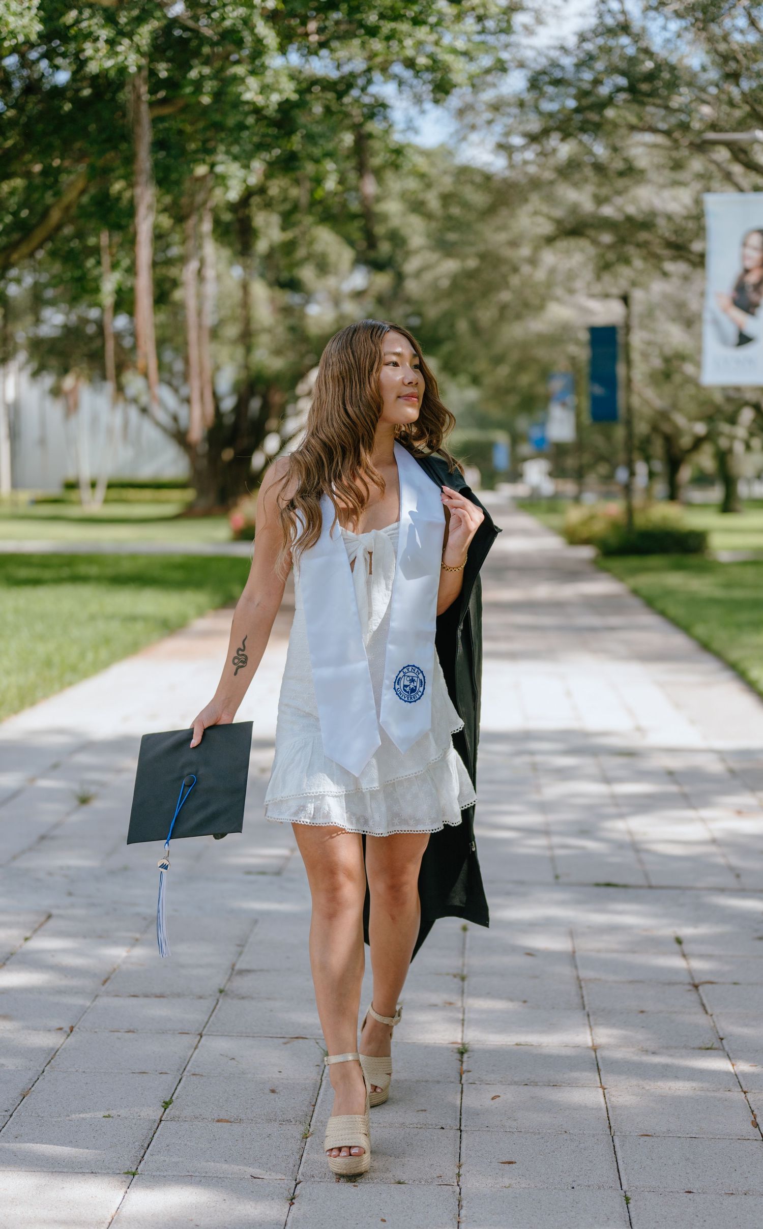 Graduate walking confidently in white dress carrying black cap and gown on a sunny campus pathway.