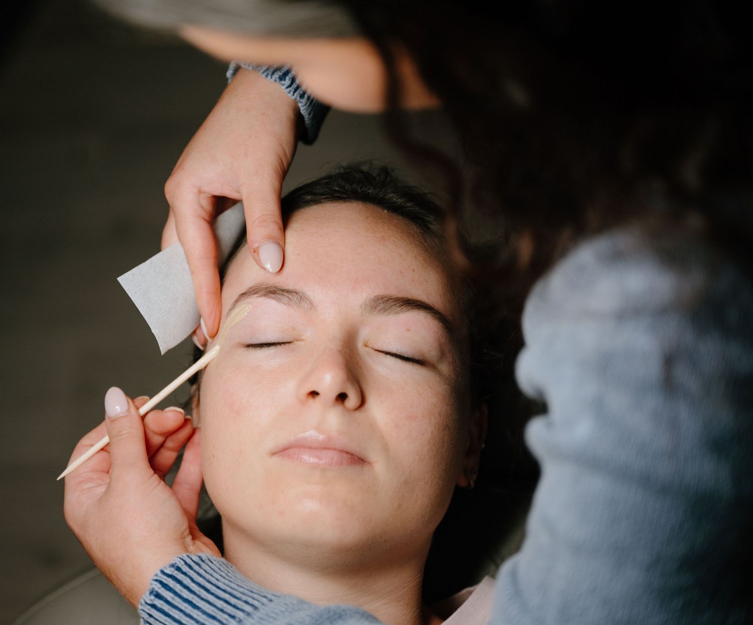 Esthetician applying wax to a young womans eyebrows during a professional brow shaping treatment.