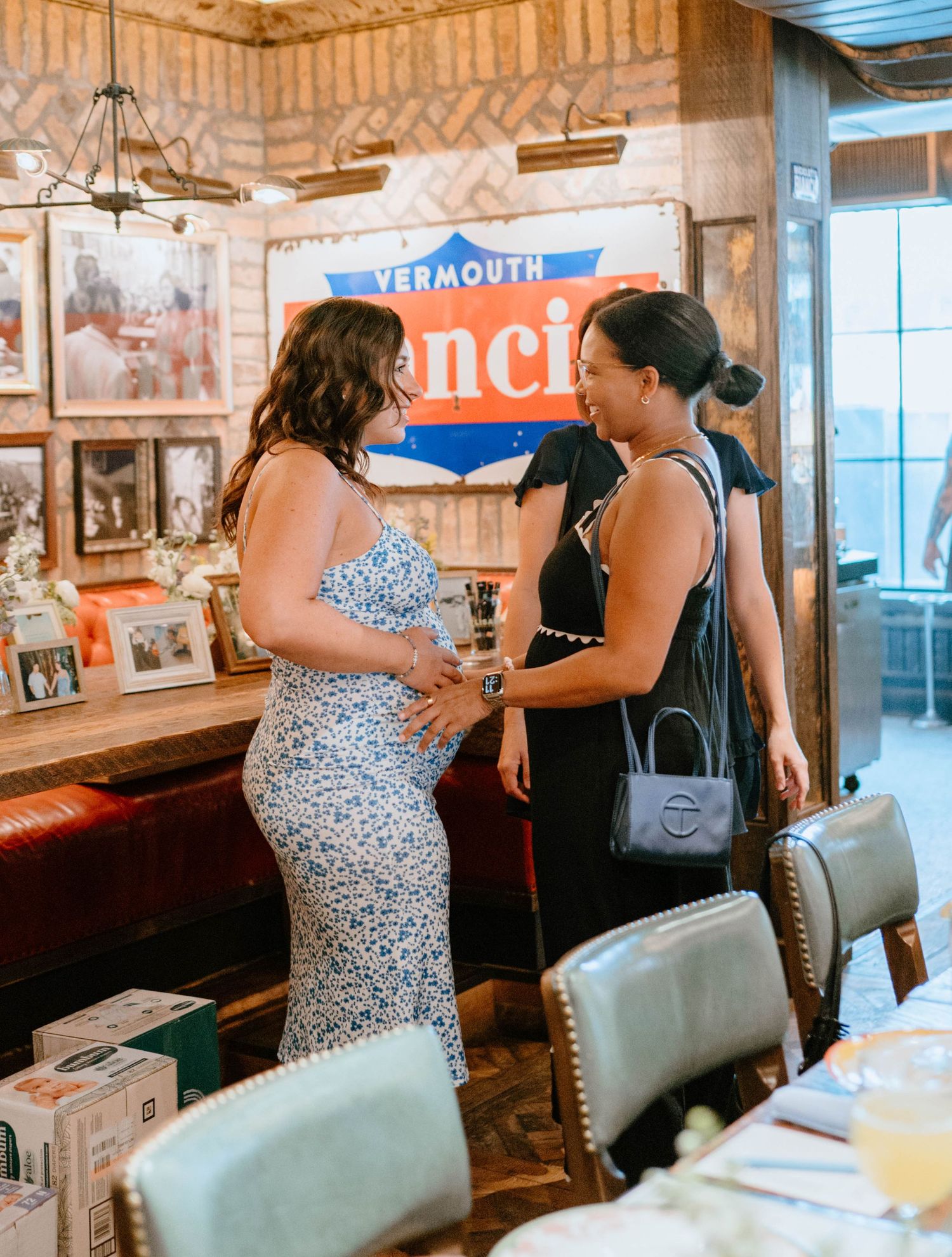 Two women in a restaurant chat warmly, one in a floral dress showing a baby bump near a vintage Vermouth sign.
