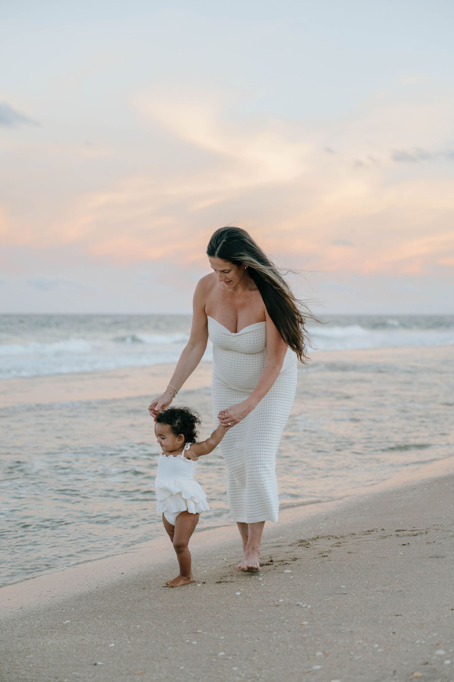 Pregnant woman in white dress walks with toddler on beach at sunset with pink clouds overhead.