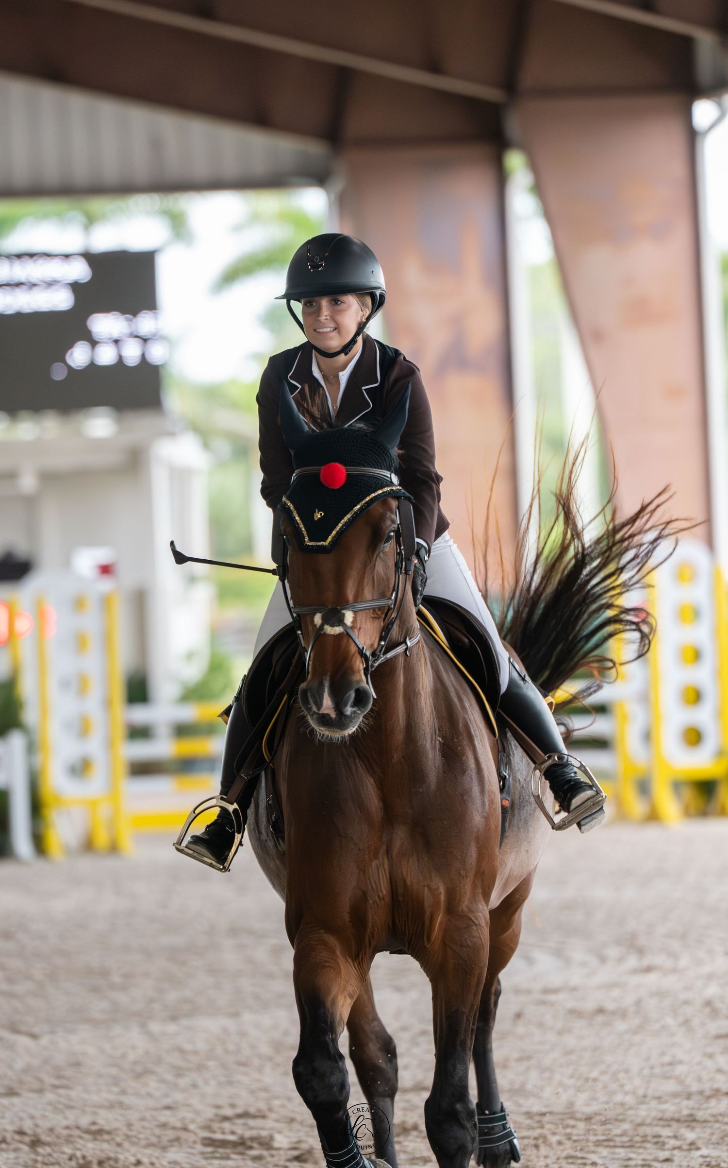 A female equestrian in black jacket and helmet rides a bay horse at an indoor show jumping arena.