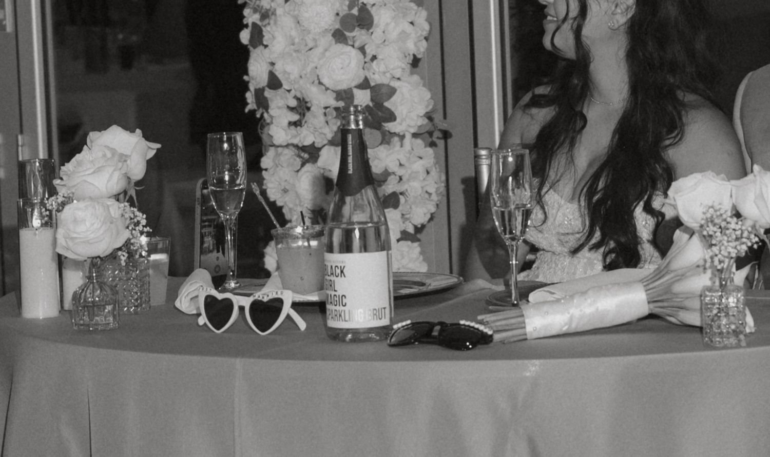 Black and white photo of wedding reception table with champagne, flowers, and heart sunglasses.