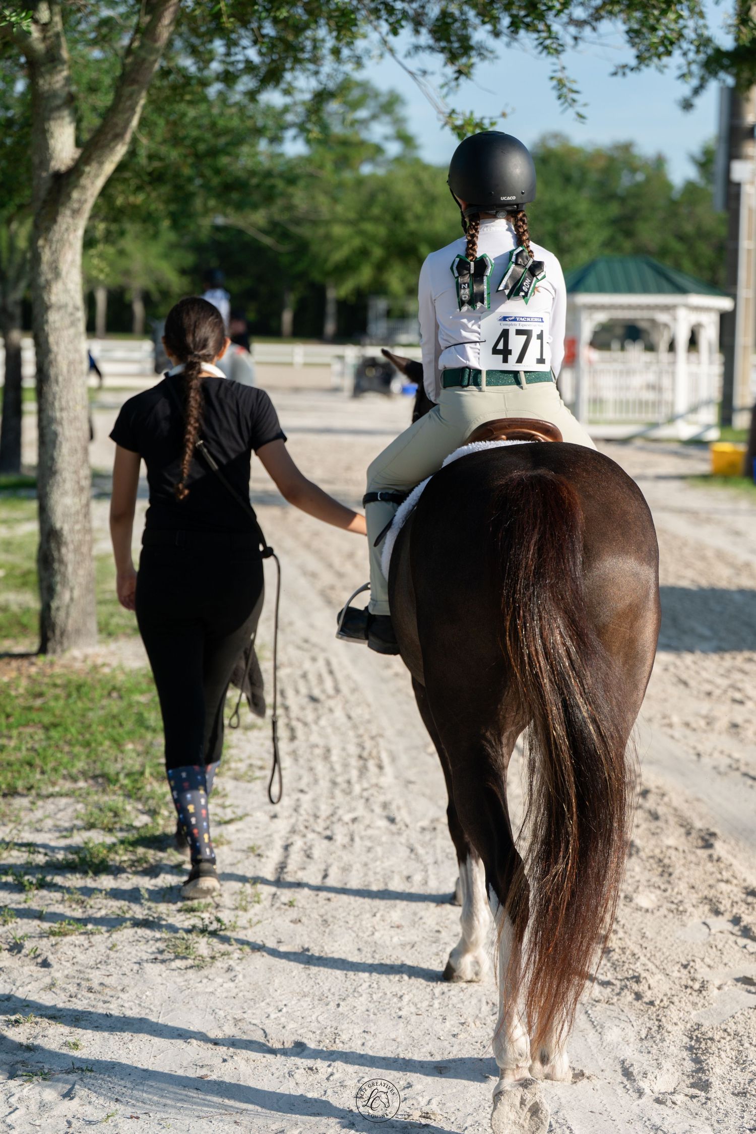 Young rider numbered 471 on chestnut horse led by trainer along sandy equestrian path.