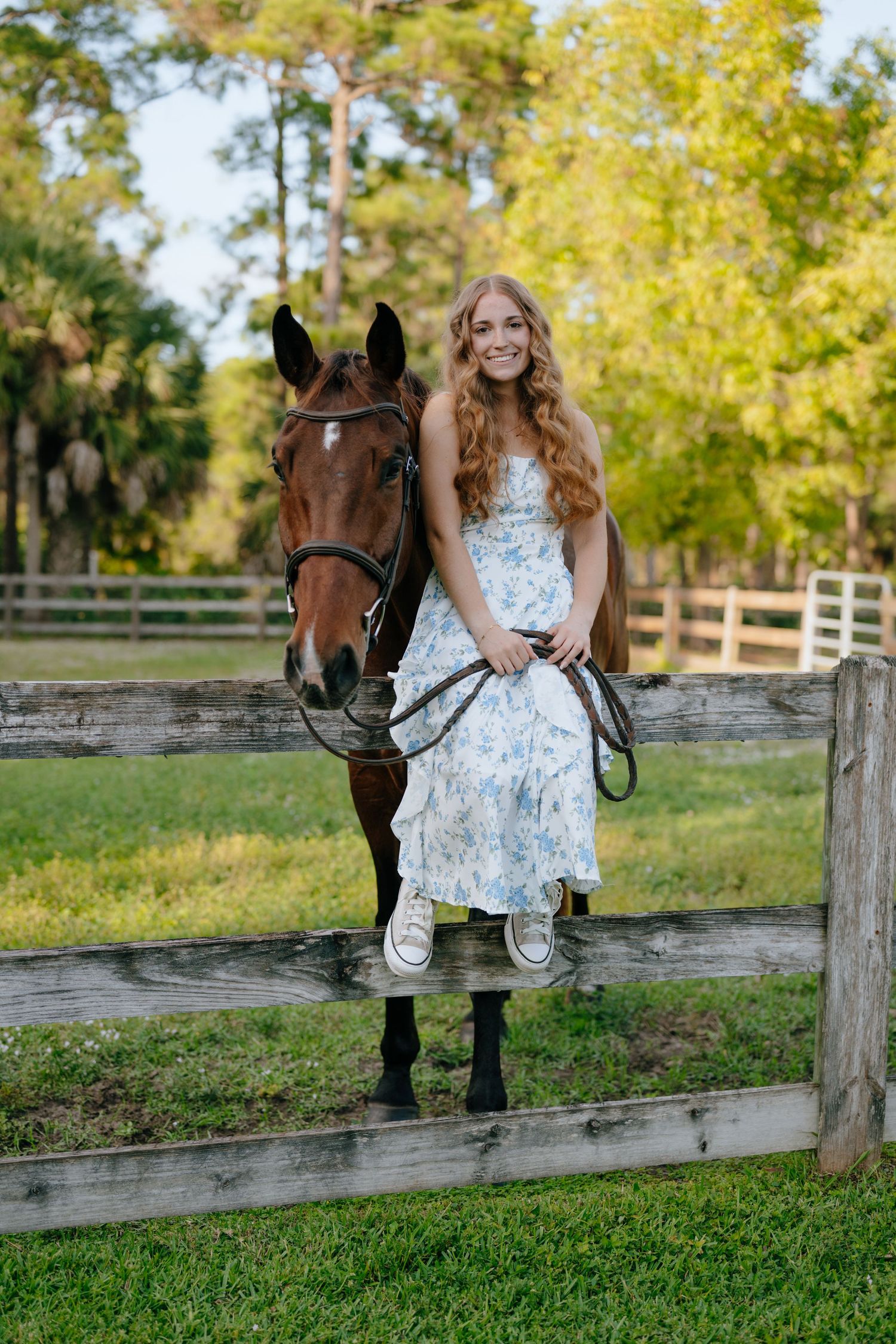 Smiling girl in floral dress sits on fence beside dark bay horse at sunny stable.