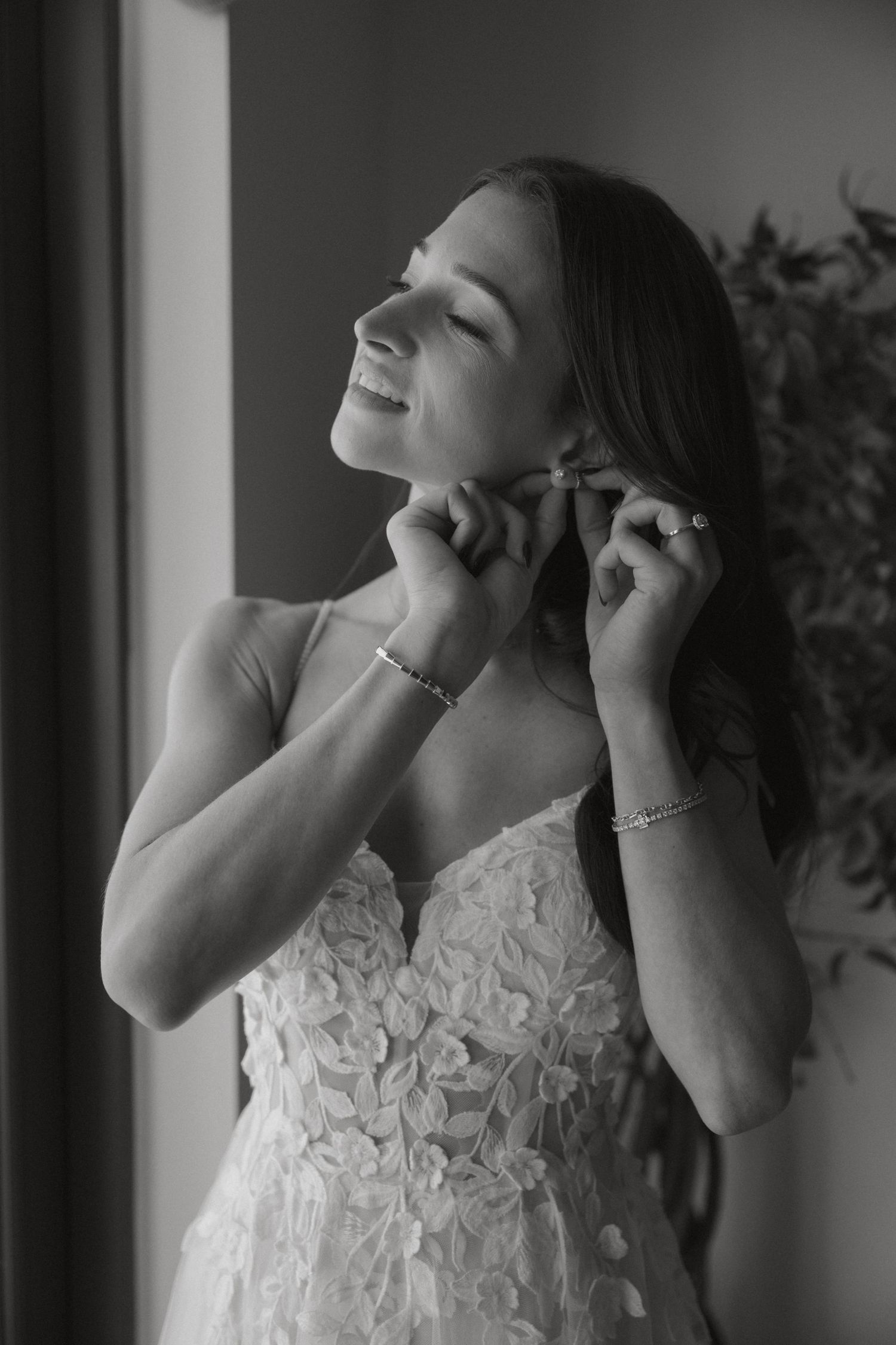 Black and white portrait of a bride in lace gown putting on earrings with a joyful expression.