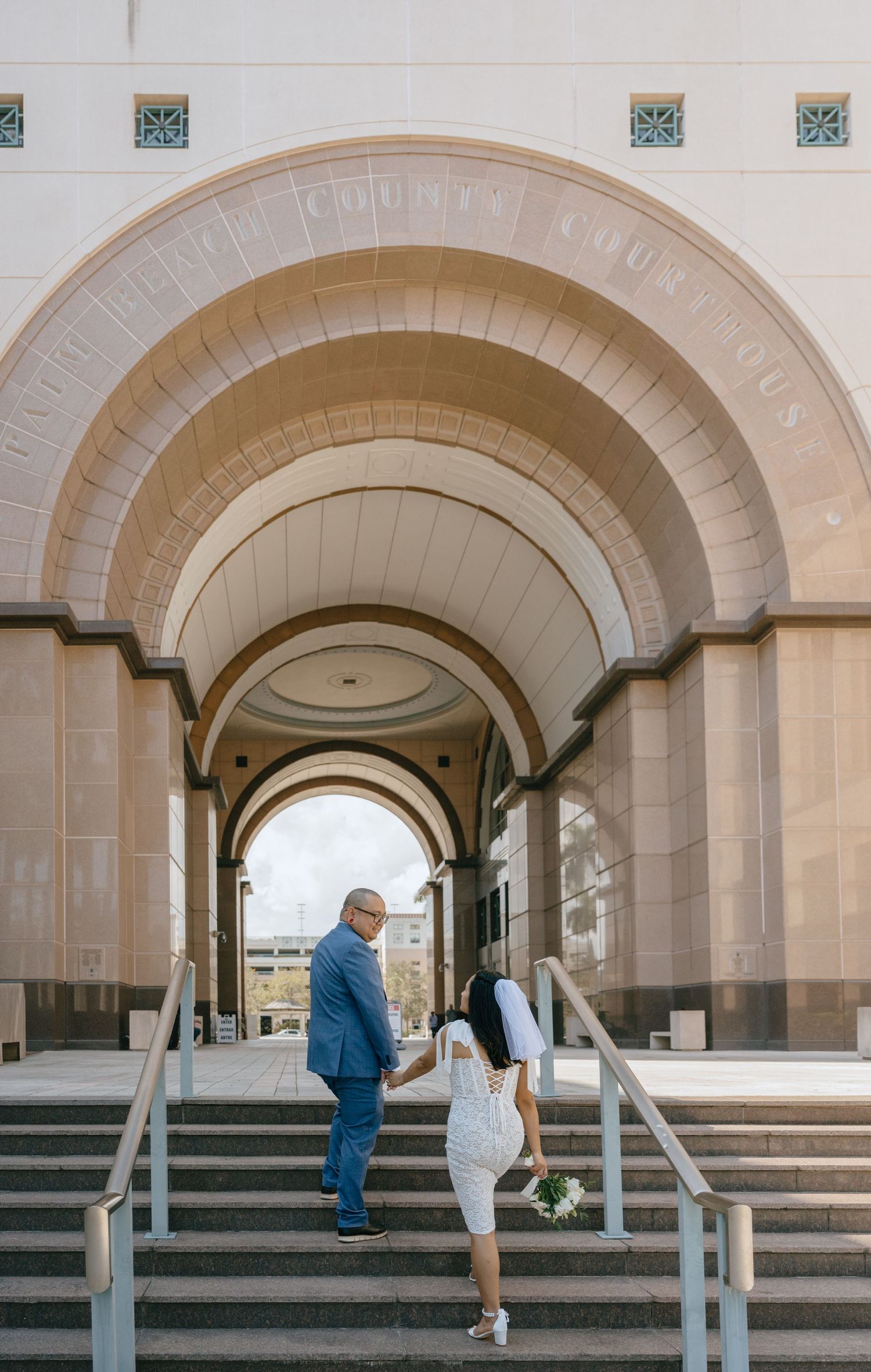 Newlywed couple holding hands walking up steps beneath grand arches of a county courthouse.