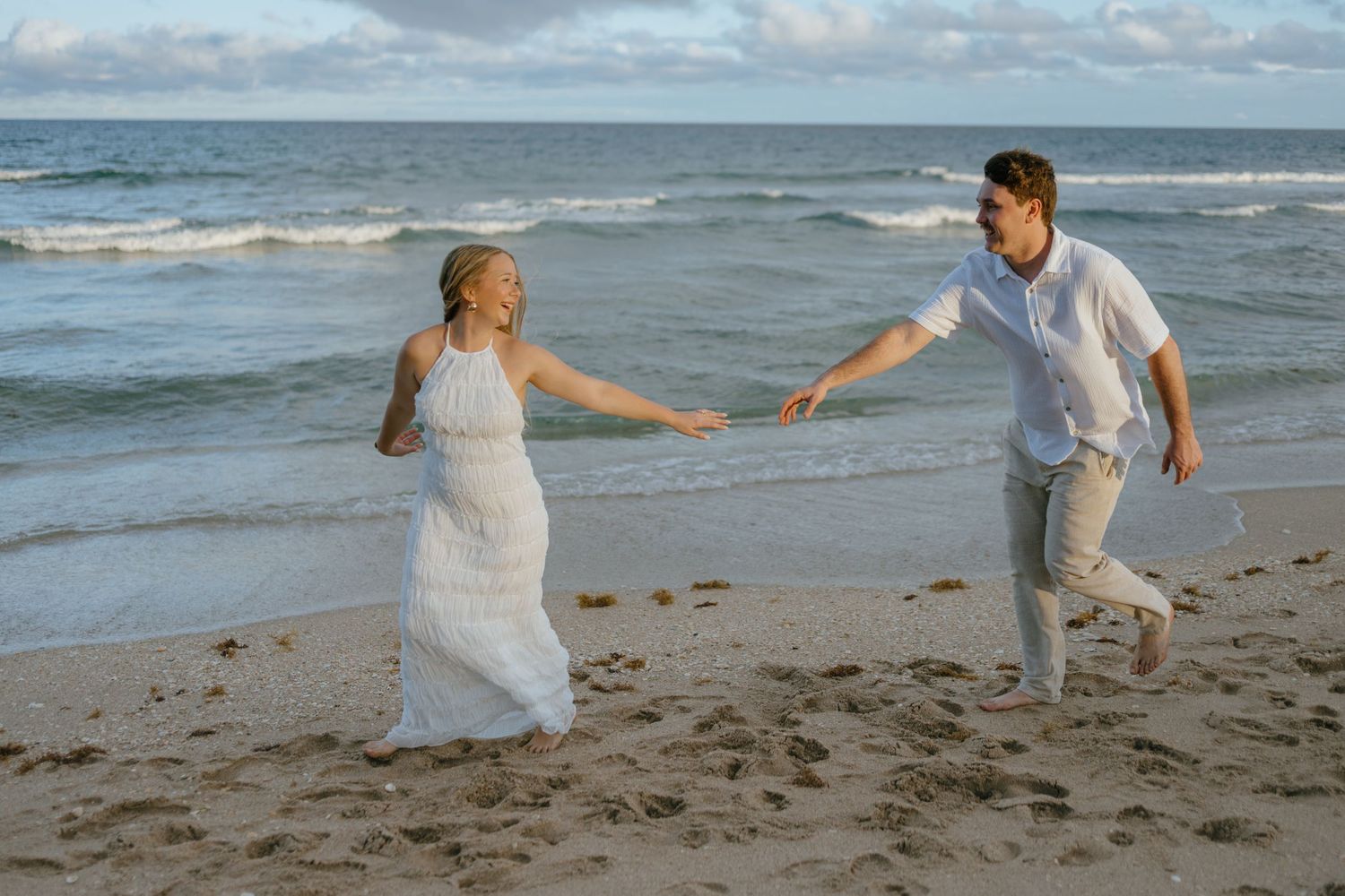 Couple in white outfits running and holding hands along a sandy beach with ocean waves in the background.
