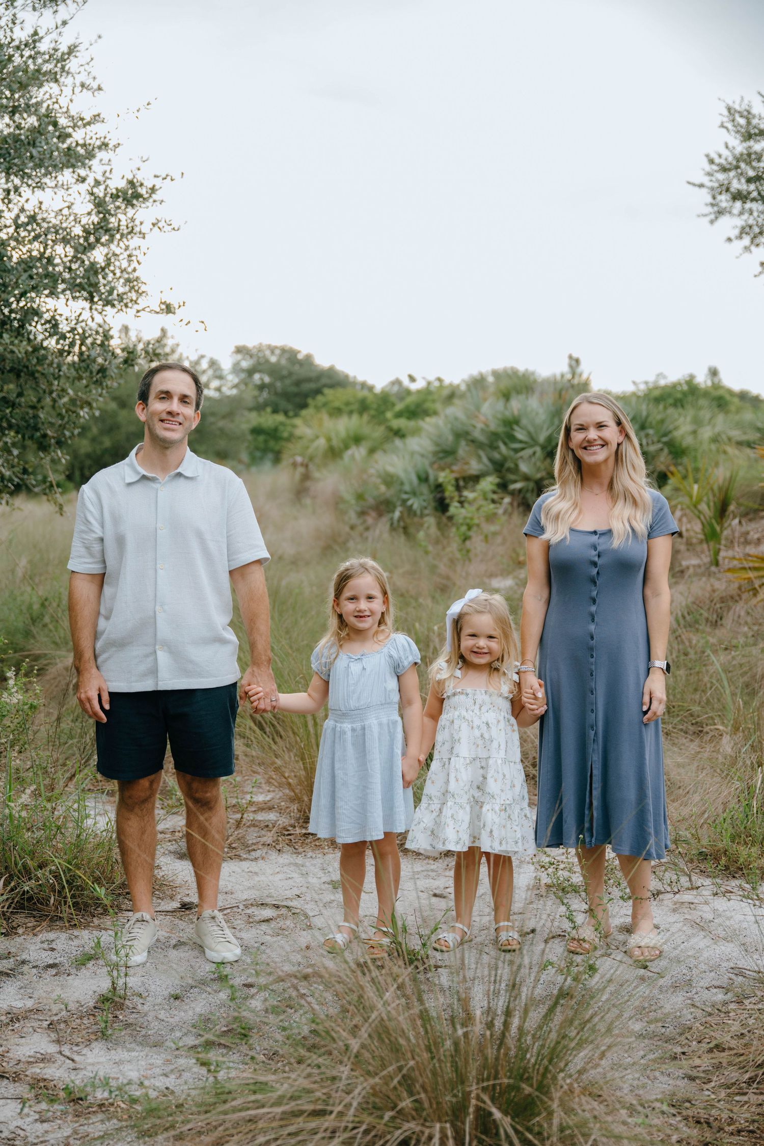 A smiling family of four poses together on a sandy path with tall grass and greenery behind them.
