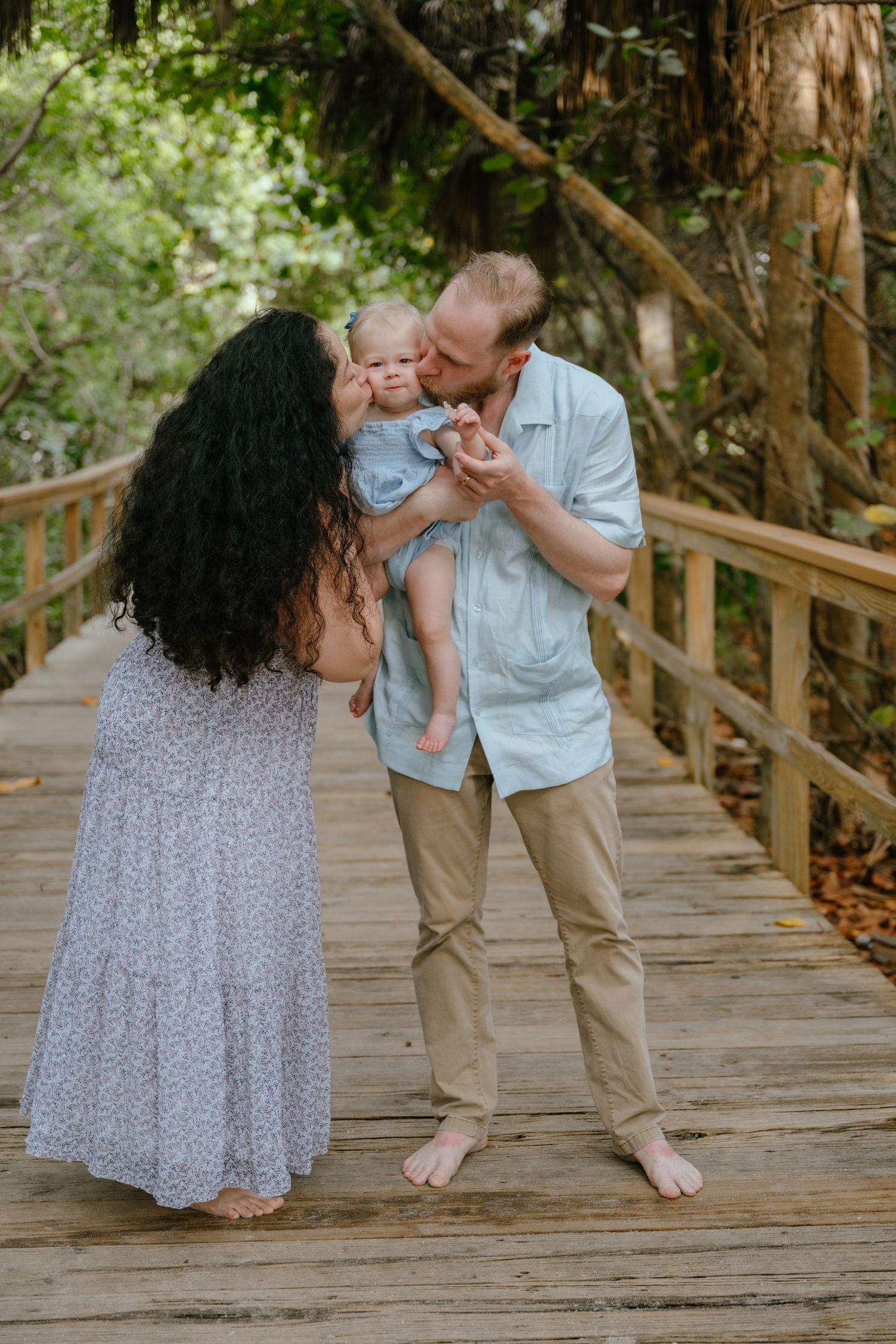 Two parents lovingly kiss their baby on a rustic wooden boardwalk surrounded by green trees.
