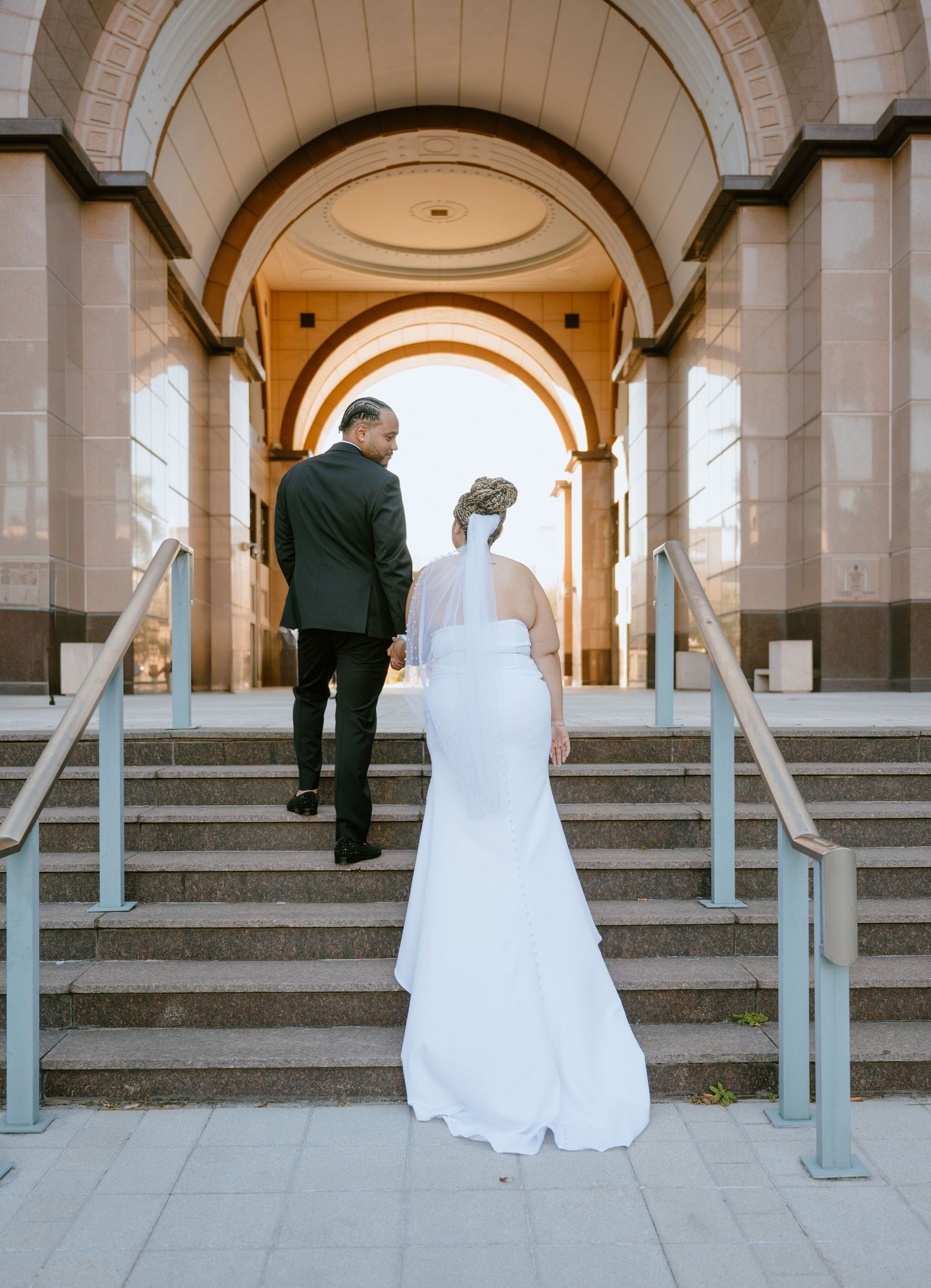 Bride in fitted white gown with veil and groom walking up steps through elegant stone arches.