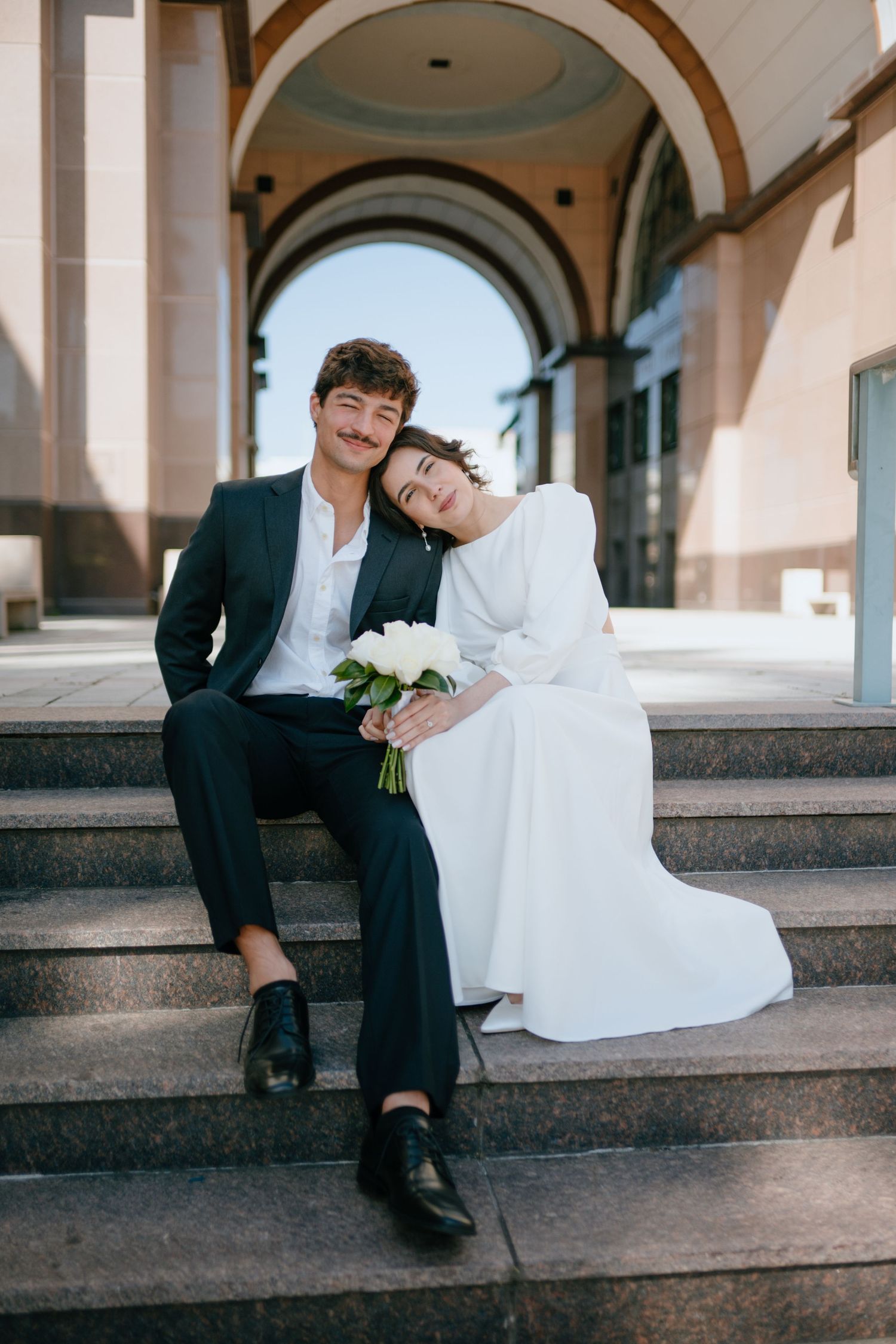 Bride resting head on groom's shoulder as they sit together on marble steps under stone arches.