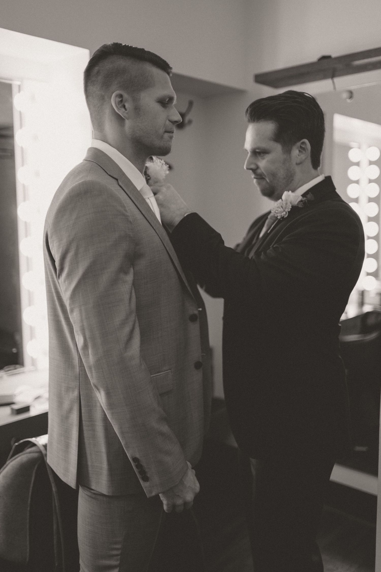 Black and white photo of groomsman pinning boutonniere on groom in suit before wedding ceremony.